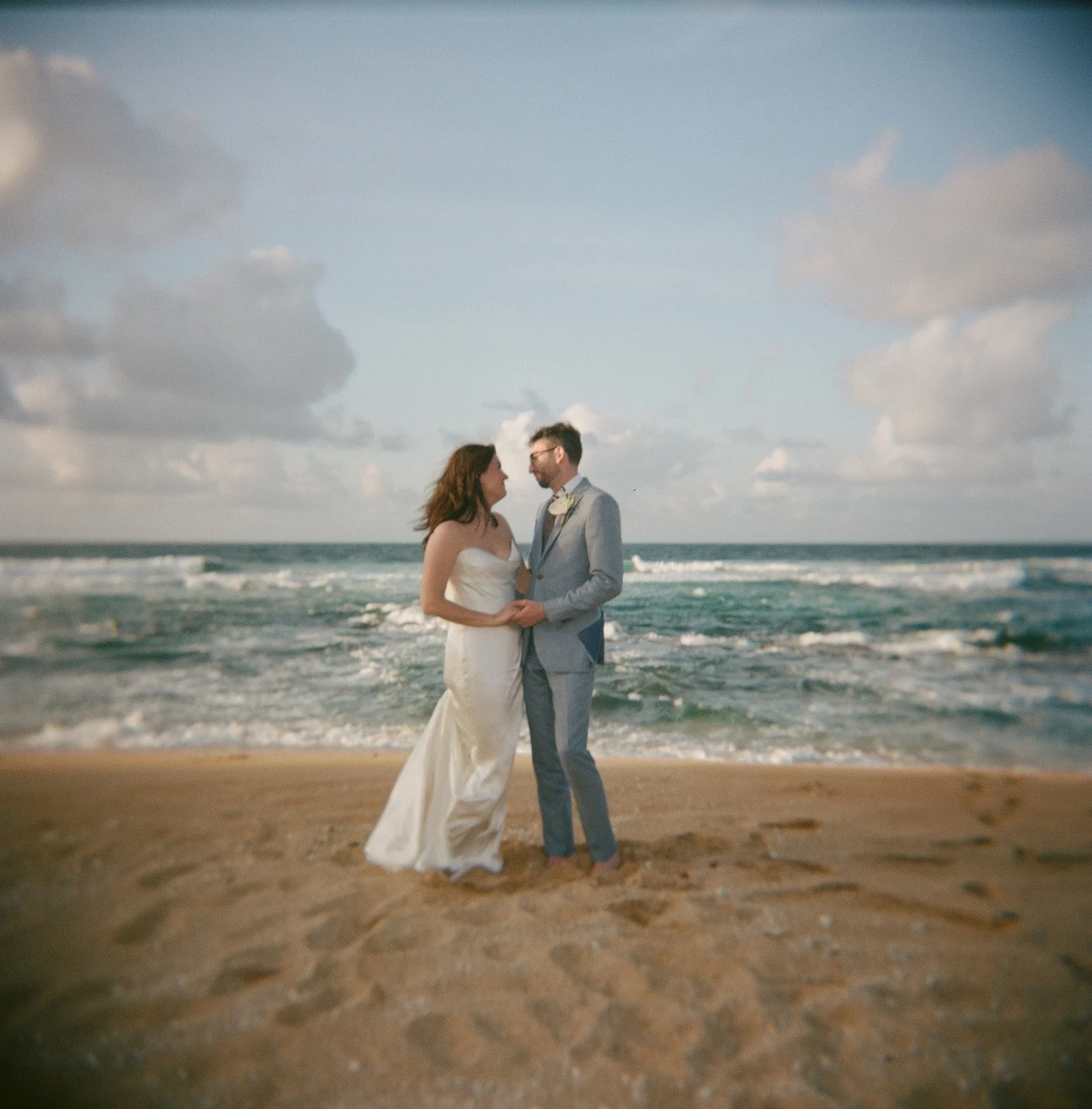 A bride and groom holding hands on a beach, looking at each other, with the ocean and cloudy sky in the background.