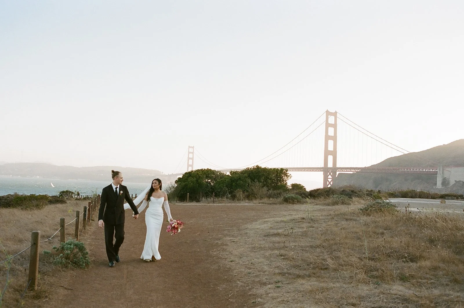 A bride and groom walking hand in hand on a dirt path near the Golden Gate Bridge with the bay in the background, during sunset or early evening.