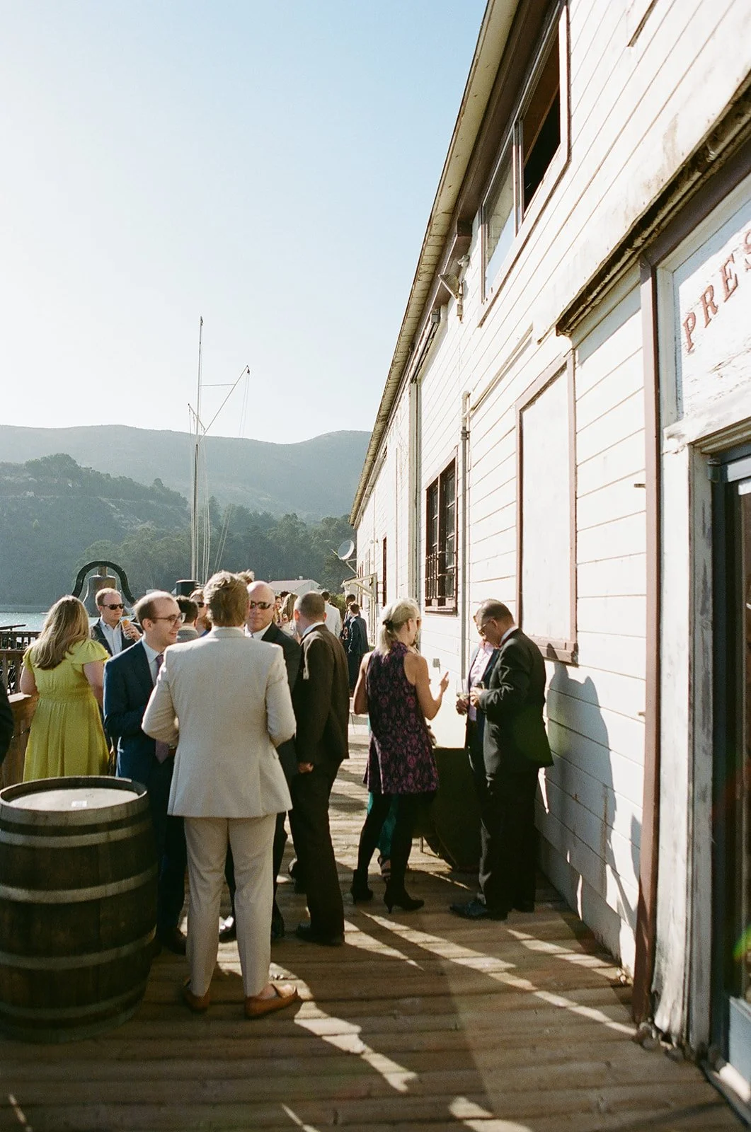 Group of people dressed in formal attire socializing outdoors near a white wooden building, with mountains and water in the background.