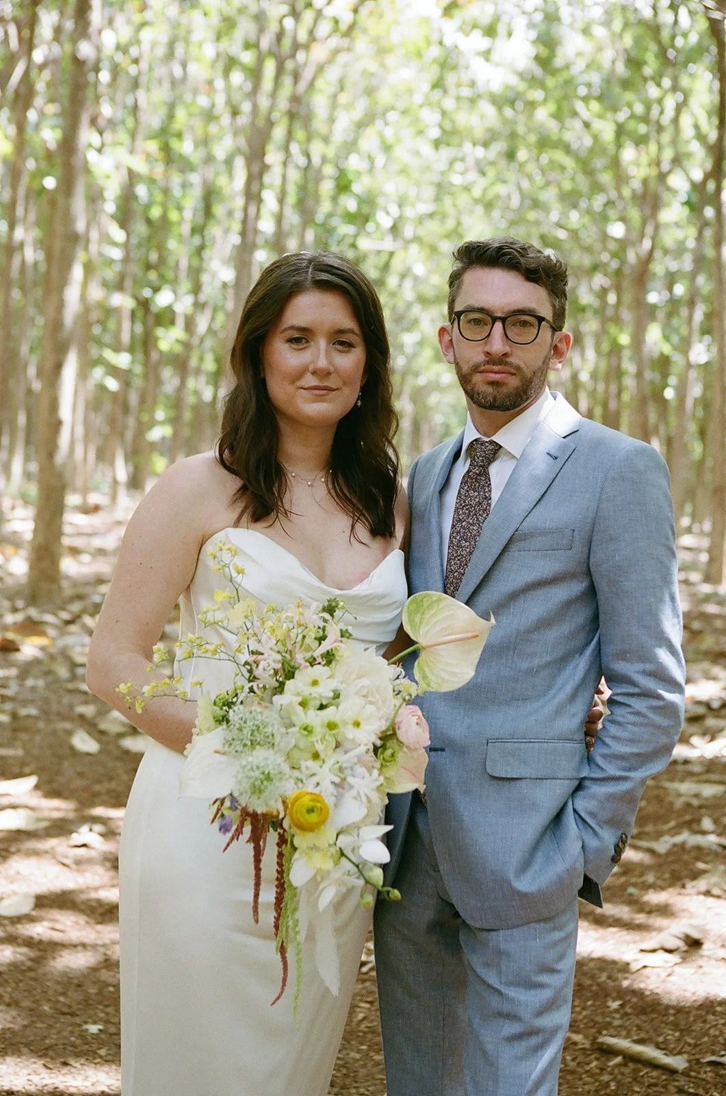 A bride and groom standing together in a wooded area for a wedding. The bride is wearing a white strapless dress and holding a bouquet of flowers. The groom is dressed in a light blue suit with a tie and glasses.