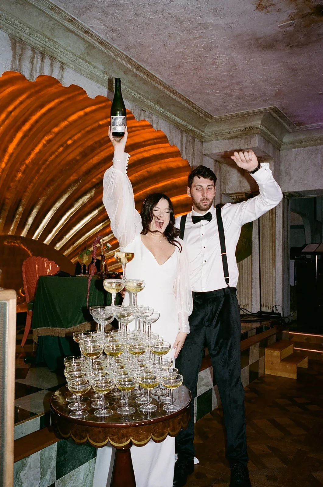 A woman in a white dress and a man in formal attire celebrating with a champagne tower at a party or wedding reception.