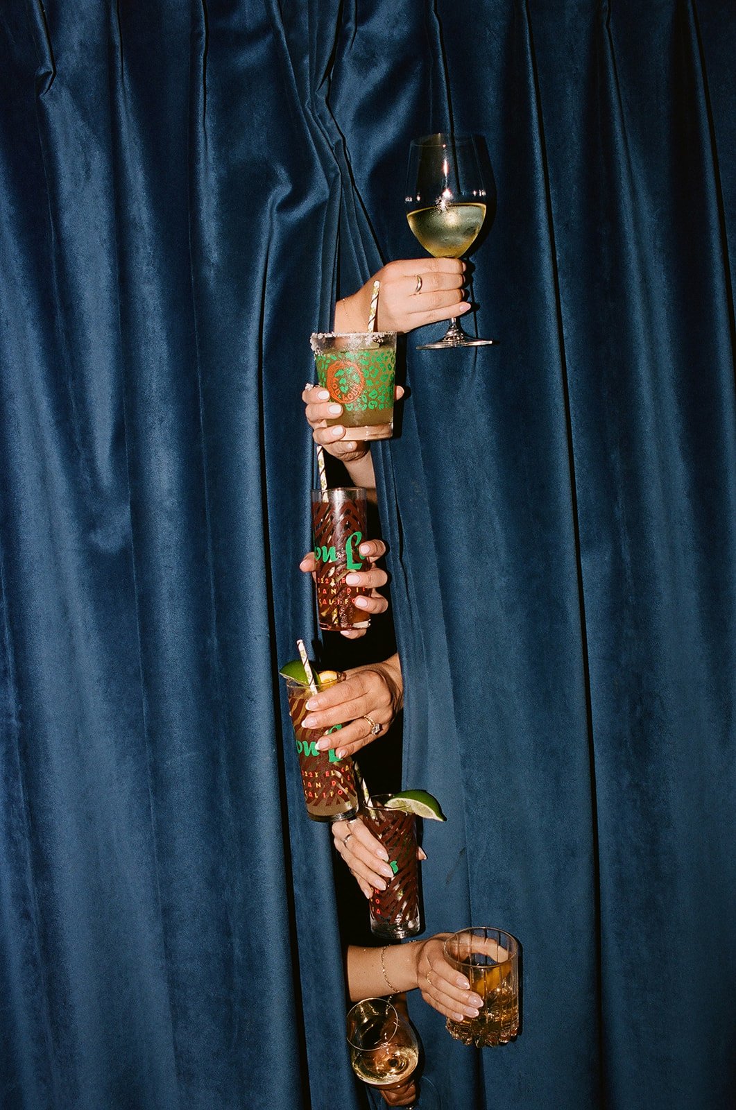 Multiple drinks held through a tear in a dark blue curtain, including wine, cocktails, and mixed drinks.