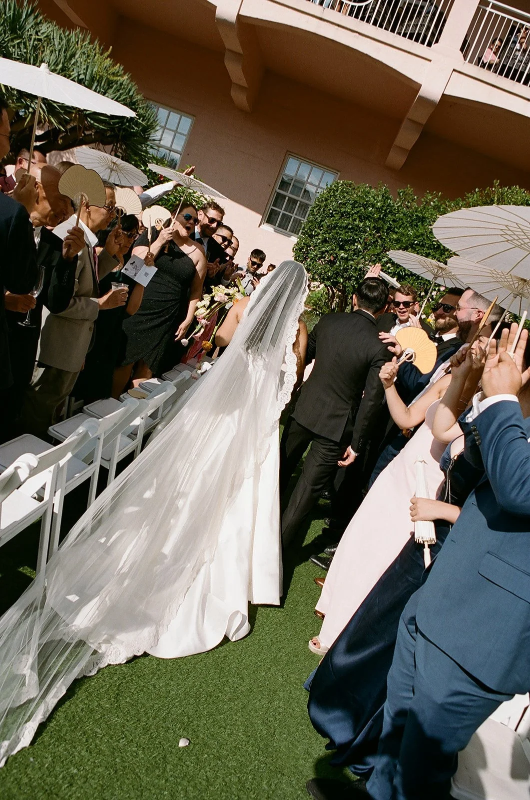 Bride in a white wedding gown and veil walking down the aisle at an outdoor wedding ceremony, with guests on both sides holding umbrellas and celebrating.