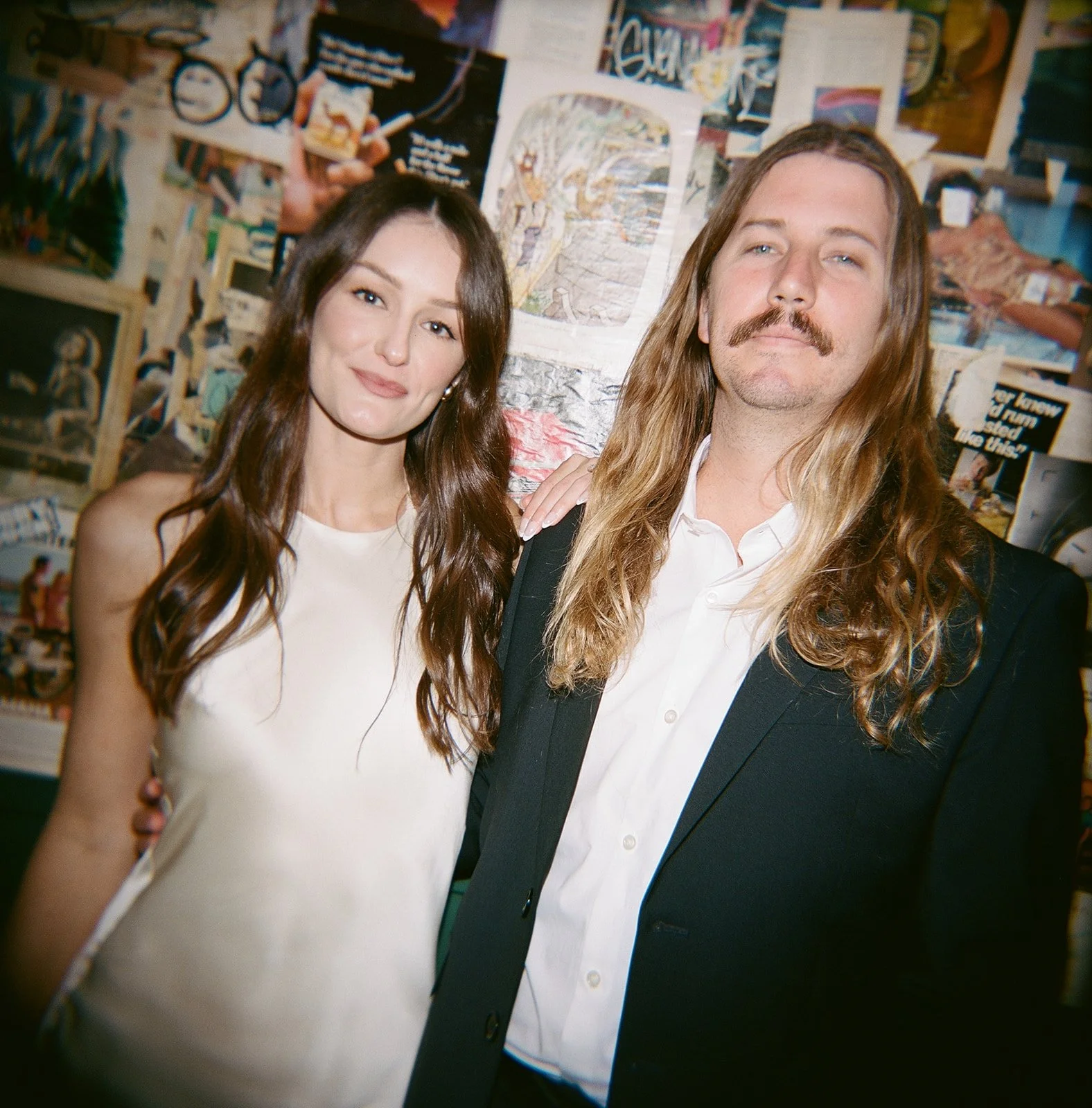 A young woman with long brown hair and a young man with long blonde hair and a mustache, standing together in front of a wall covered with artwork and posters, smiling at the camera.