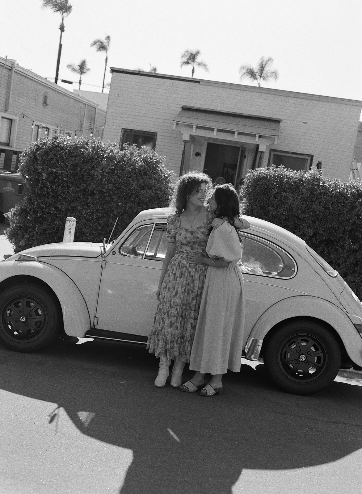 Two women standing close and smiling in front of a classic car on a sunny street, with houses and palm trees in the background.