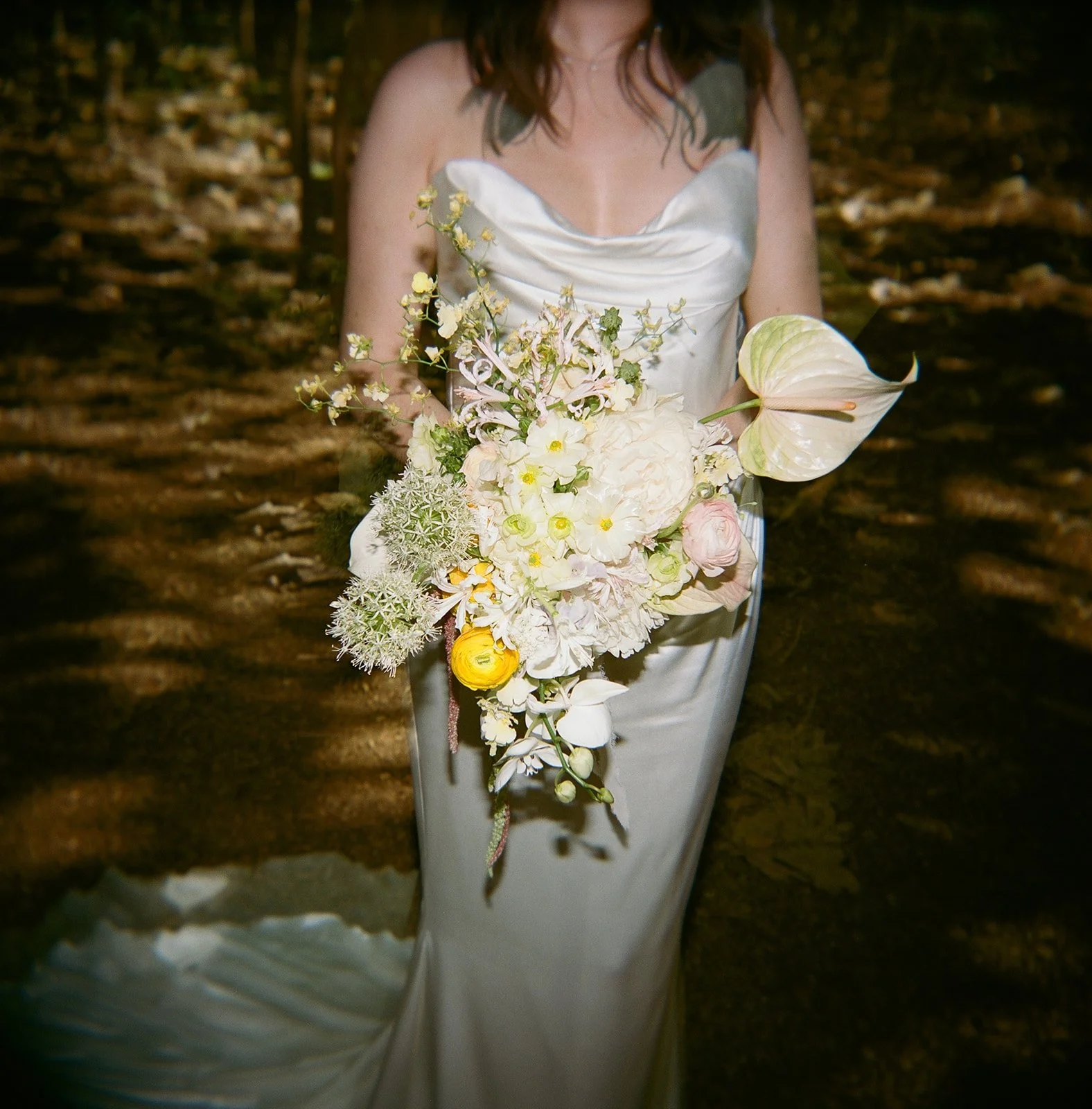 Woman in a satin dress holding a cascading bouquet of white and pale yellow flowers with green foliage, set outdoors on a wooded trail.