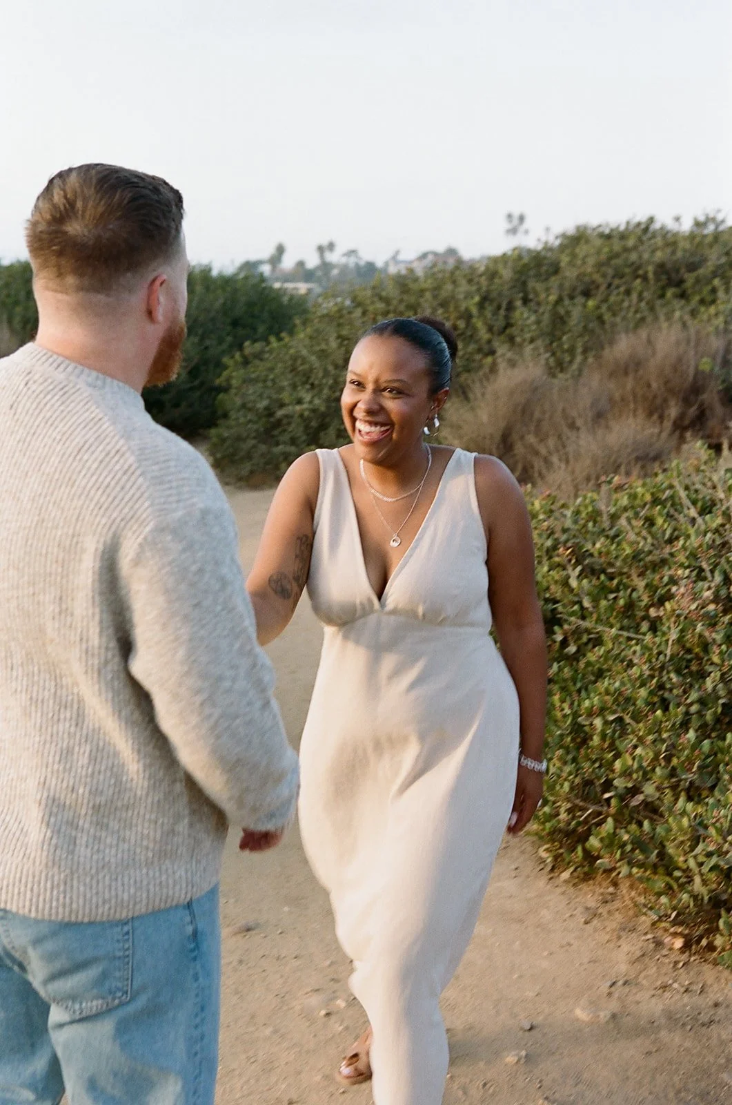 A woman in a cream-colored dress smiling and reaching out to a man holding her hand outdoors.