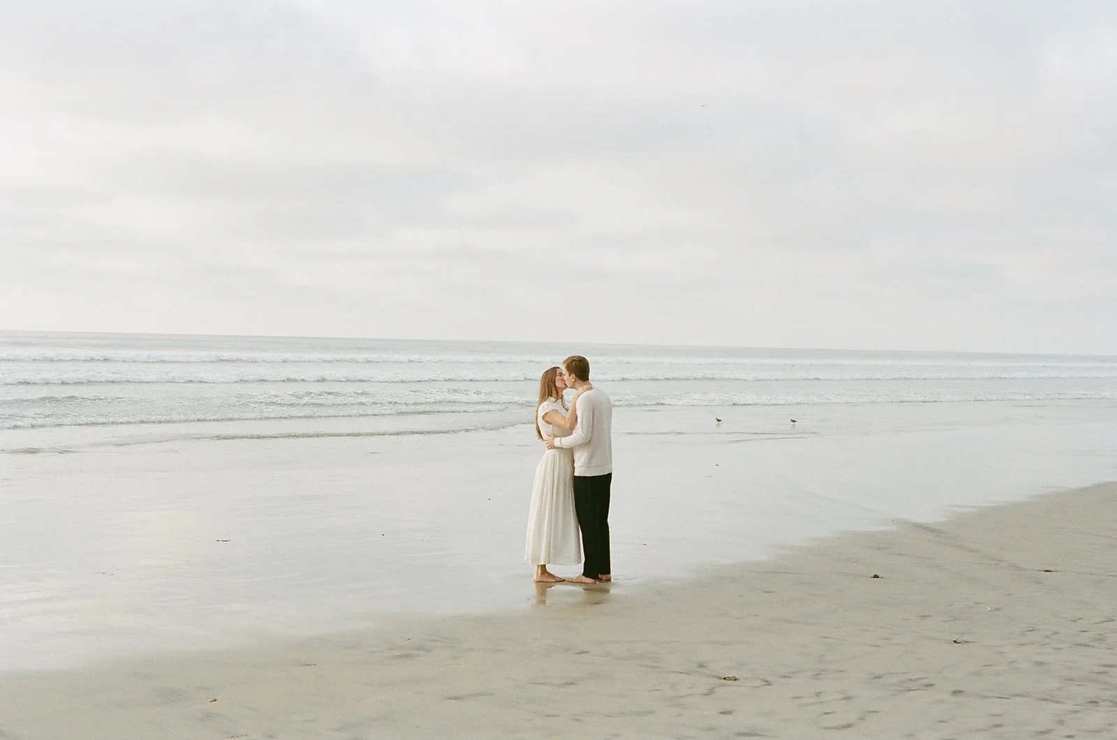 A couple dressed in white standing and kissing on a beach with gentle waves and an overcast sky.