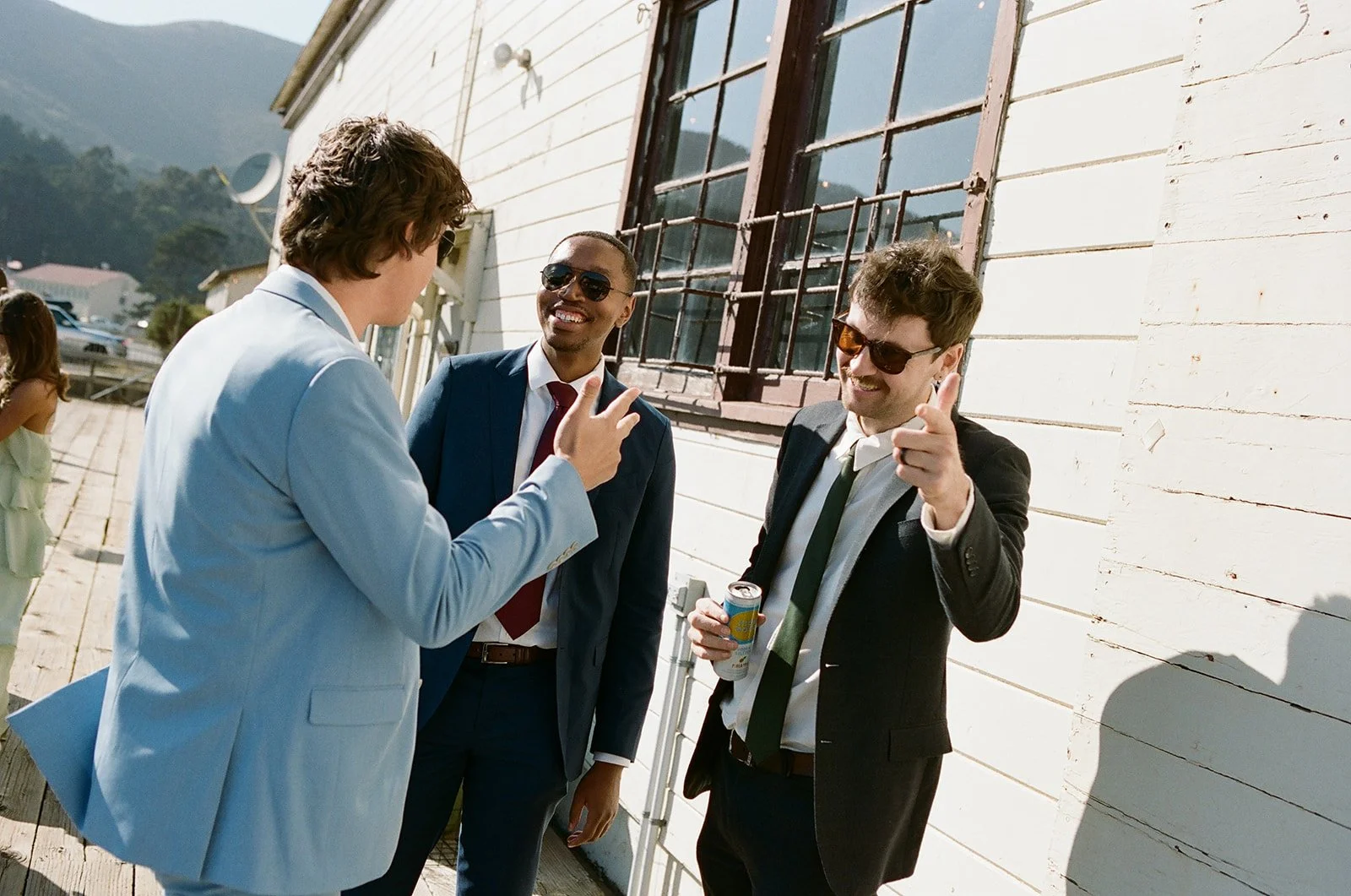 Three men in suits having a conversation outdoors near a white wooden building, with mountains in the background. One man is pointing and smiling, holding a soda can. Another man is gesturing with his hand, and the third man is smiling and wearing su