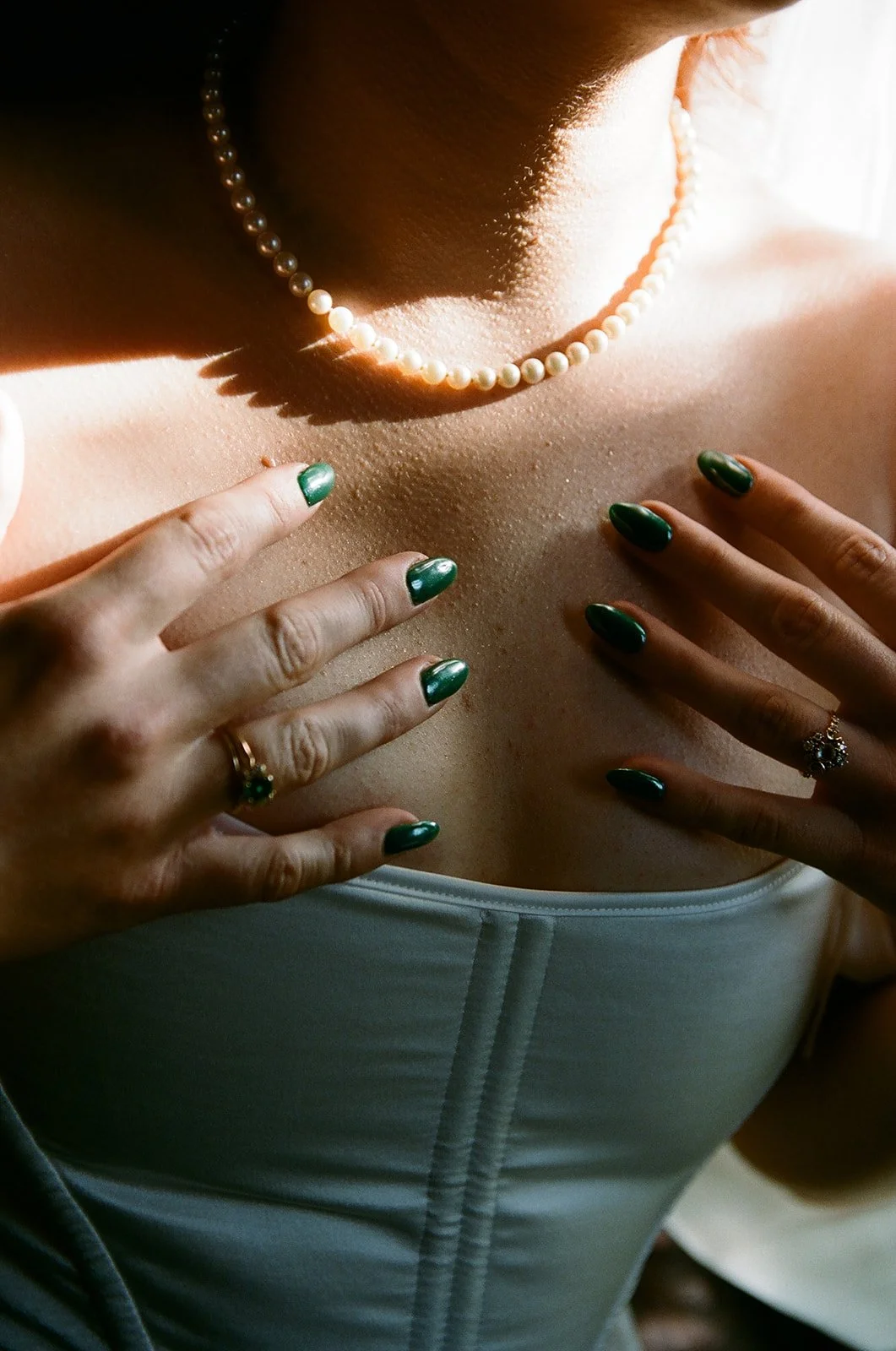 A woman wearing a pearl necklace and jewelry, with green nail polish, touching her chest near her collarbone.