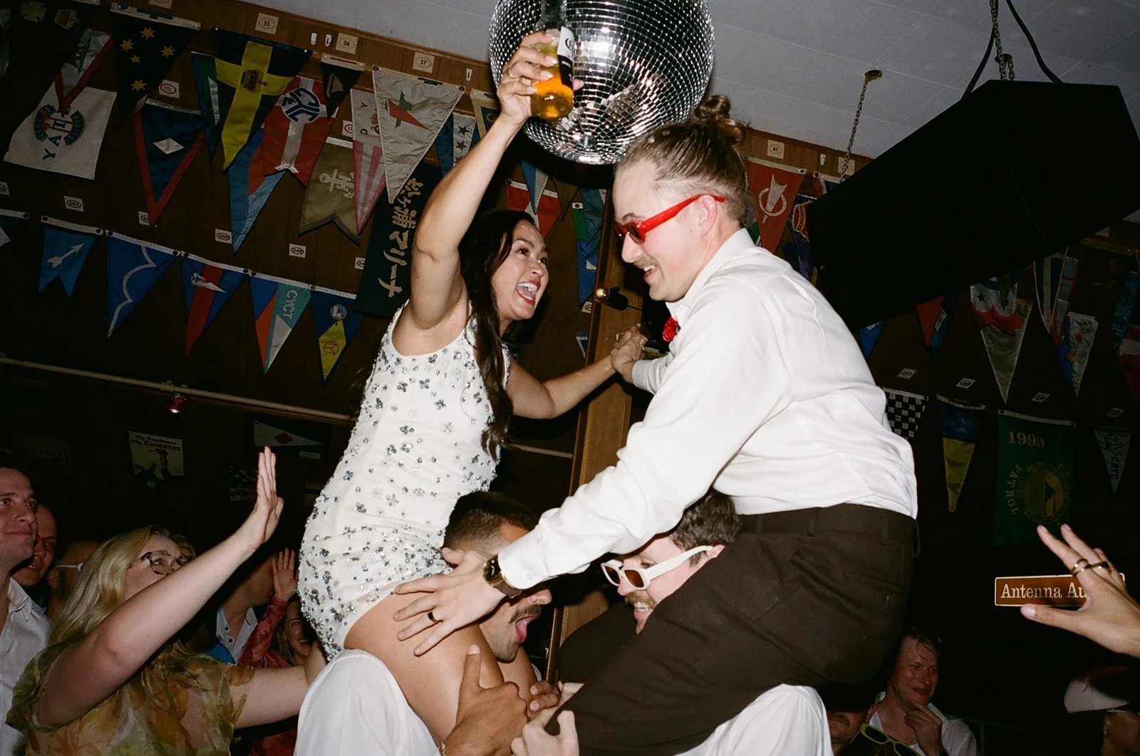 People celebrating at a lively party, one woman sitting on a man's shoulders holding a beer, with a disco ball overhead and colorful flags in the background.