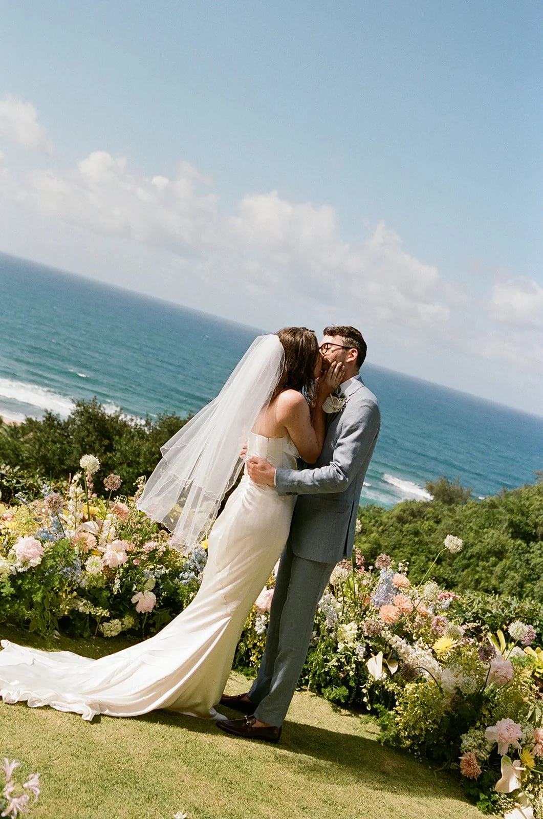 A bride and groom kissing outdoors near the ocean with a floral arrangement in the foreground.