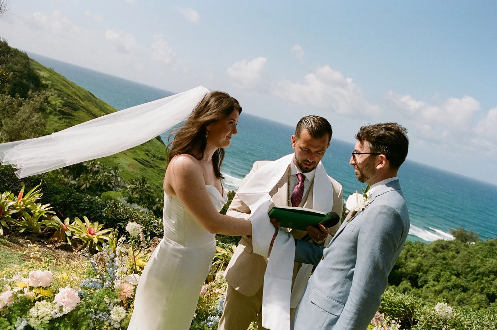 A wedding ceremony taking place outdoors near the coast, with a bride, groom, and officiant in front of an ocean and green hillside, surrounded by colorful flowers.