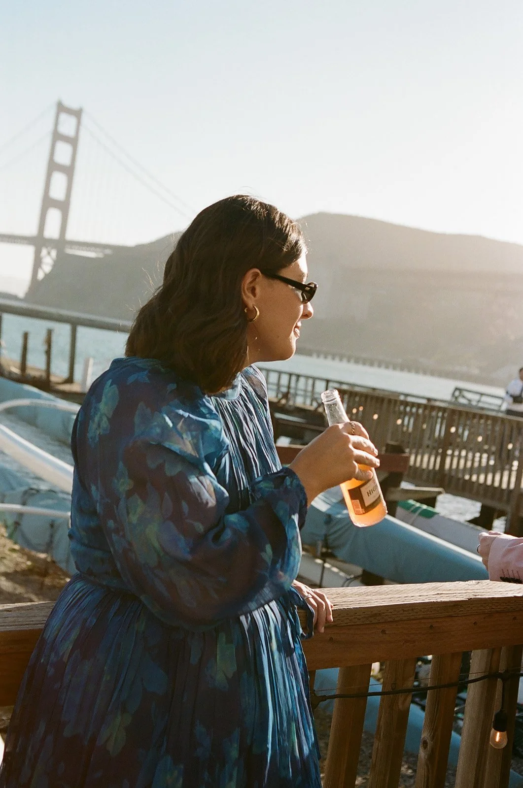 A woman with dark hair, wearing sunglasses and a blue dress, smiling and holding a bottle, standing on a wooden deck overlooking water and a bridge.