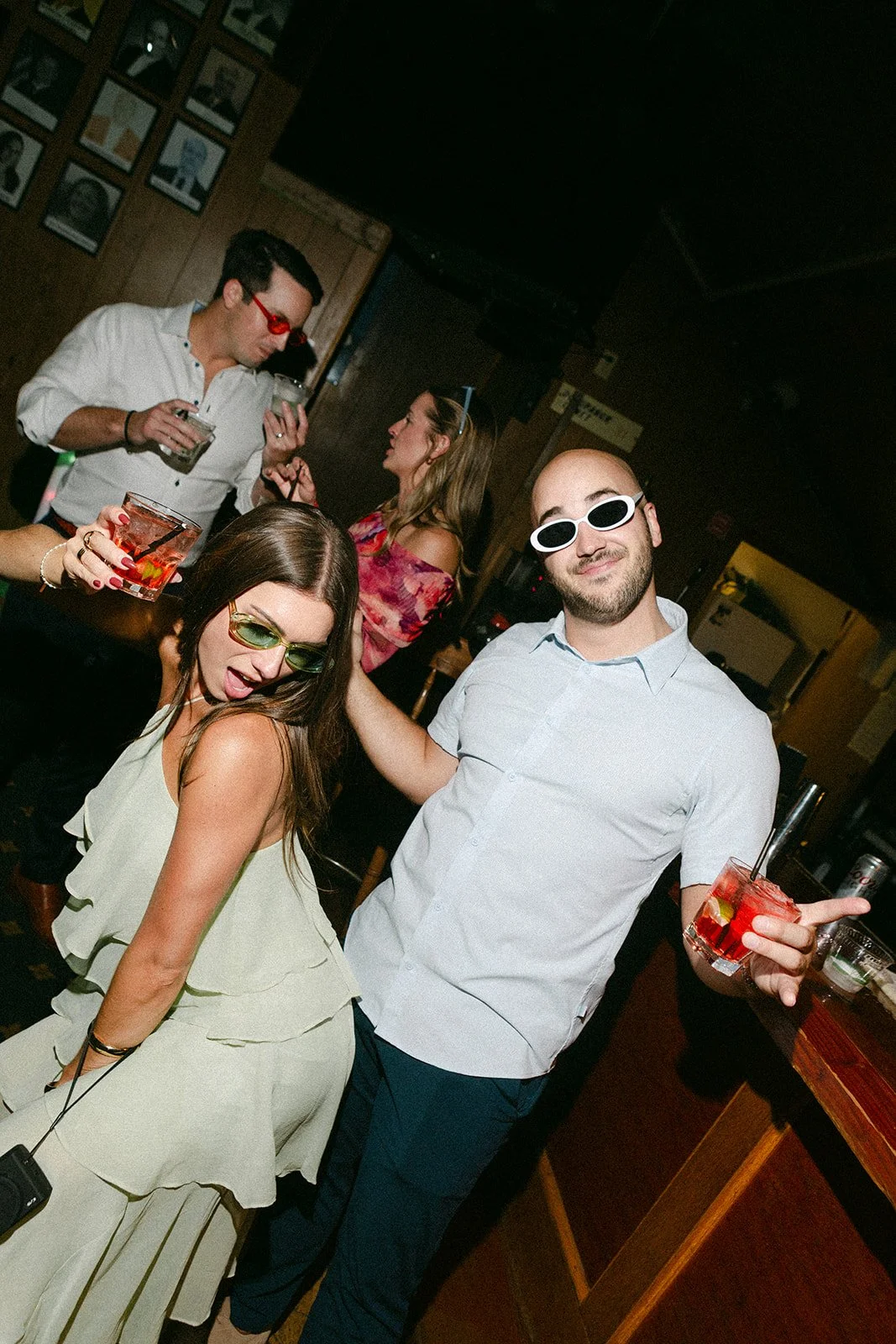 People partying at a bar, wearing sunglasses, holding drinks, dancing, and socializing.