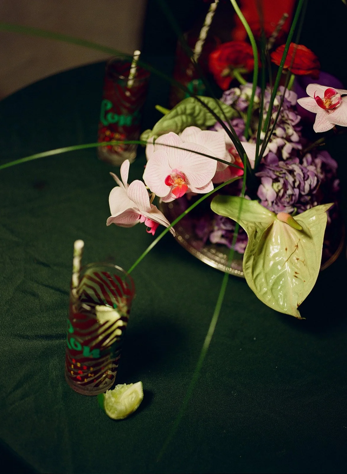 A floral arrangement of pink orchids, purple flowers, and red blossoms with green leaves in a metallic bowl. Two striped glasses with drinks and straws are in front on a dark table, along with a lime wedge.