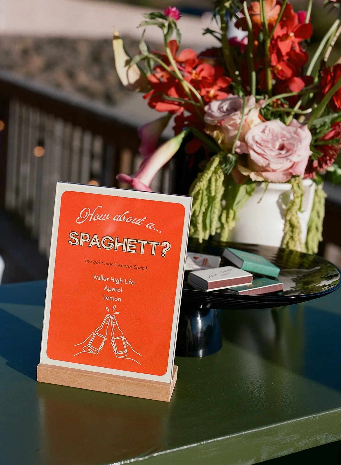 A sign on a green table reads, 'How about a... SPAGHETT? The poor man's Aperol Spritz! Miller High Life, Aperol, Lemon,' with a drawing of two beer bottles clinking. Behind the sign, there is a black tray with some small boxes and snacks. A large bou