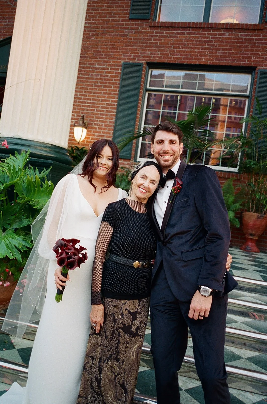 A wedding celebration with four people standing outside in front of a brick building, including a bride in a white dress holding a bouquet of dark red calla lilies, a groom in a tuxedo, an older woman in a black dress with gold patterns, and a man in