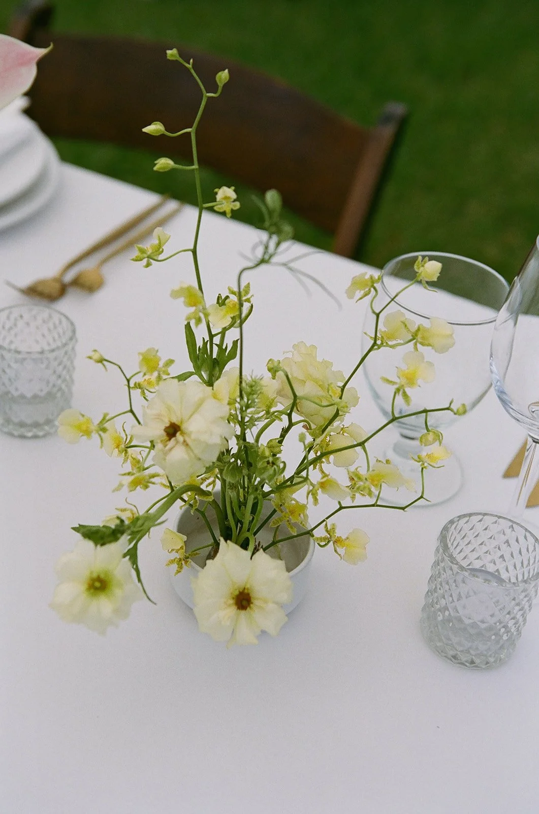 A white flower arrangement in a white vase on a table set with clear textured glassware and white tablecloth.