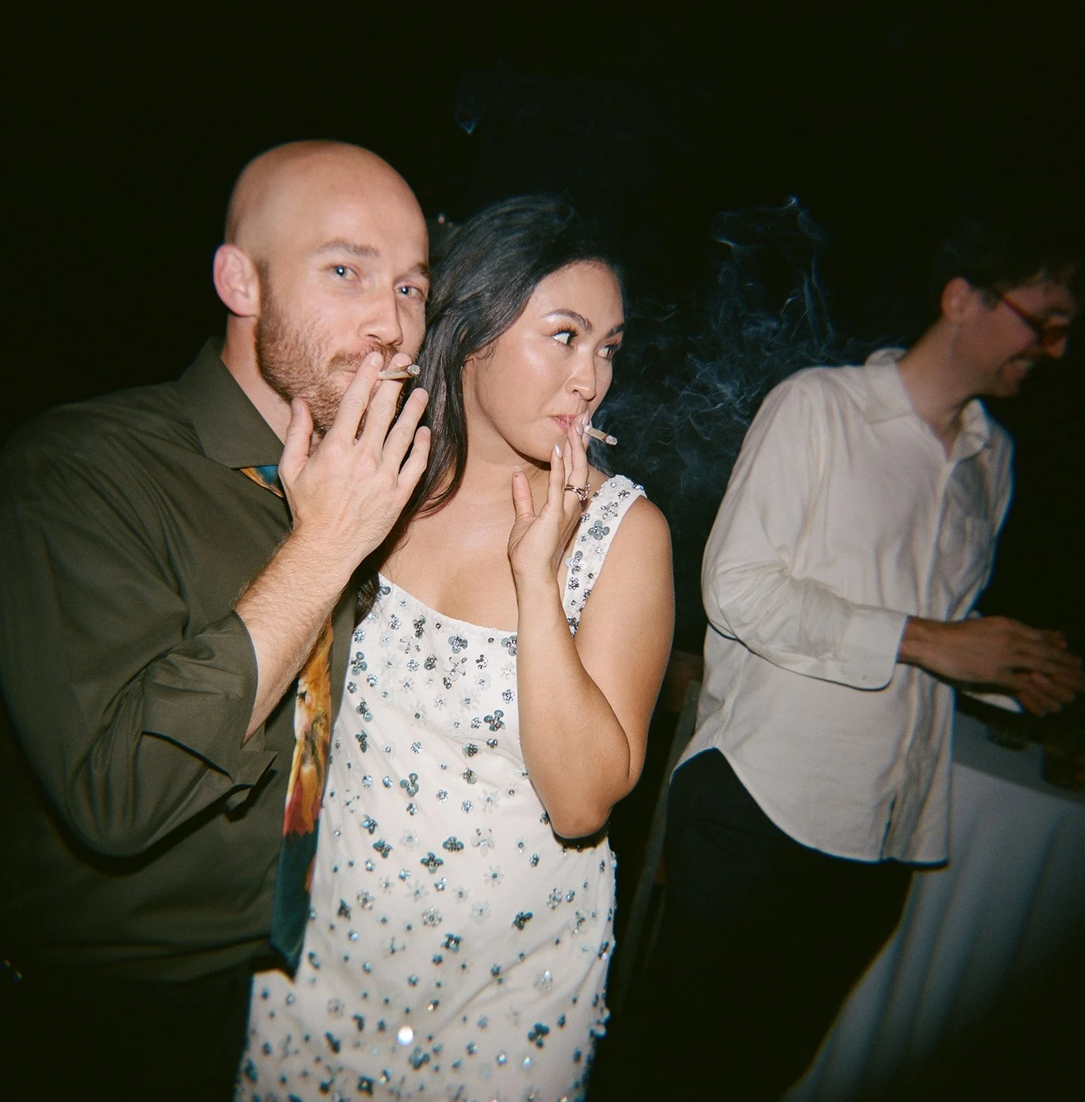 Two people smoking cigarettes at a party with a third person nearby.