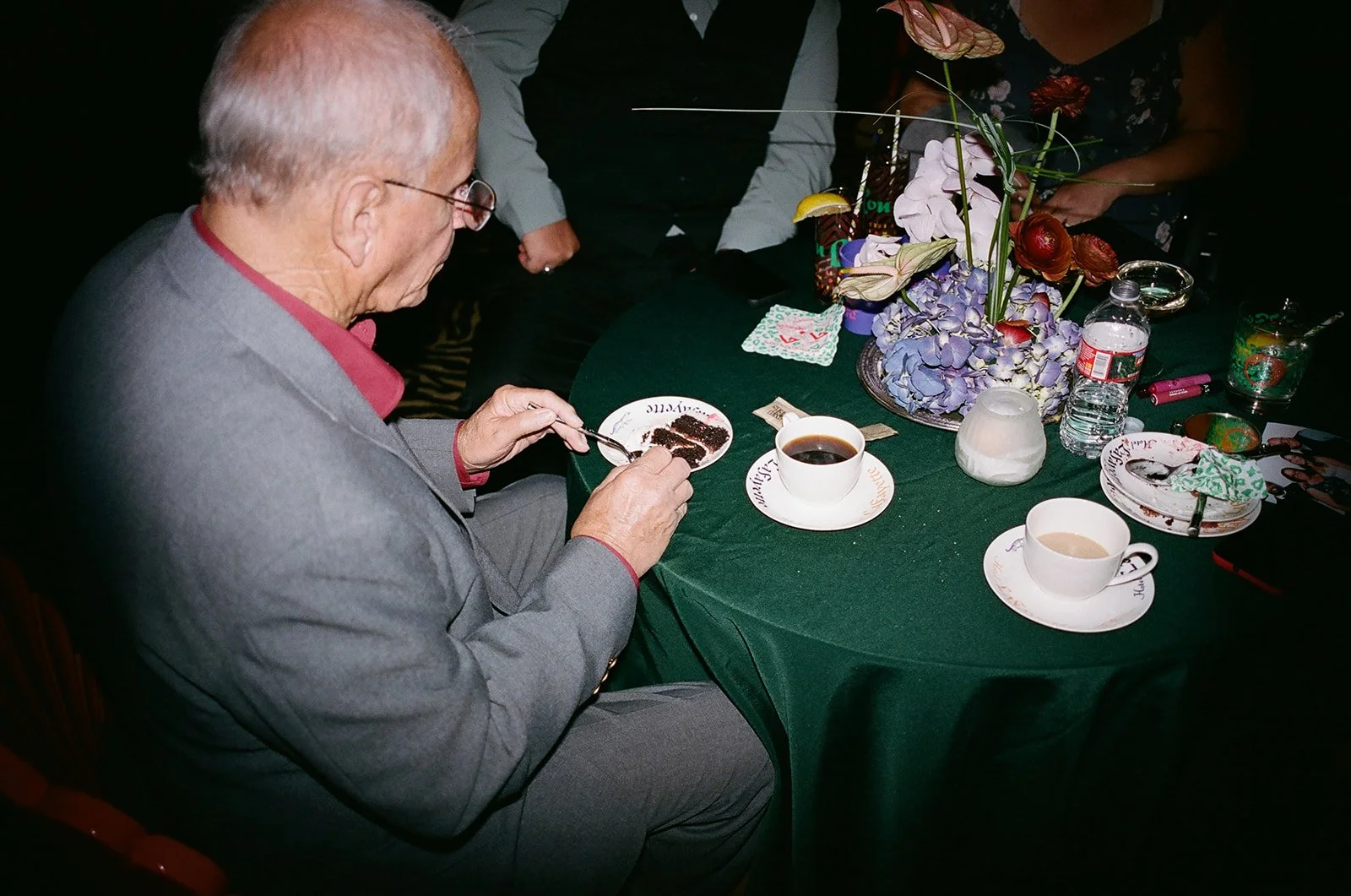 An elderly man in a gray suit and red shirt is sitting at a green table, carefully serving himself a slice of chocolate cake with a fork. The table is decorated with a large floral centerpiece featuring purple and red flowers. There are cups of coffe