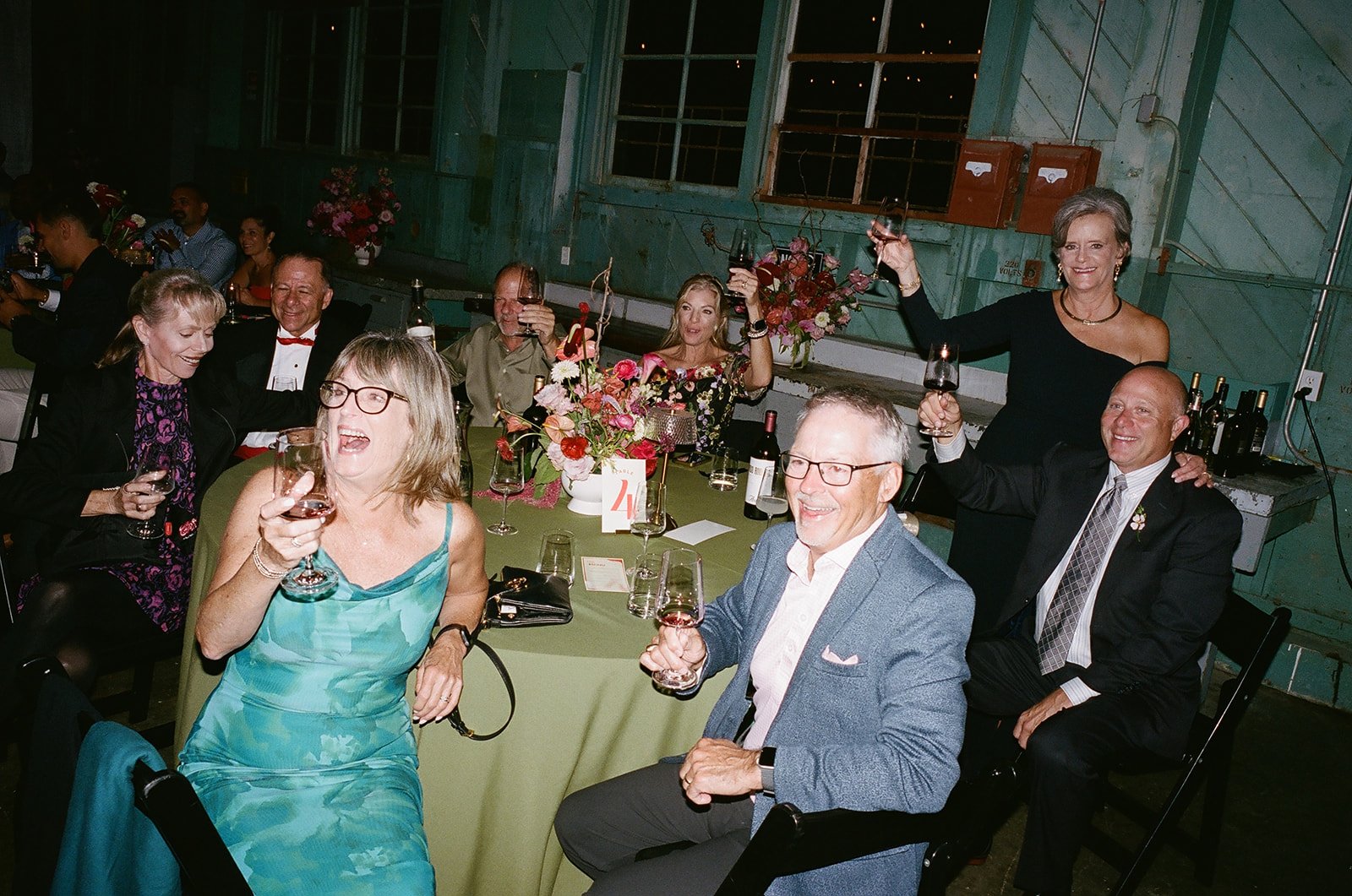 People at a celebration or dinner party raising glasses of wine, sitting around a table with floral centerpieces, inside a rustic, green-painted barn or hall.
