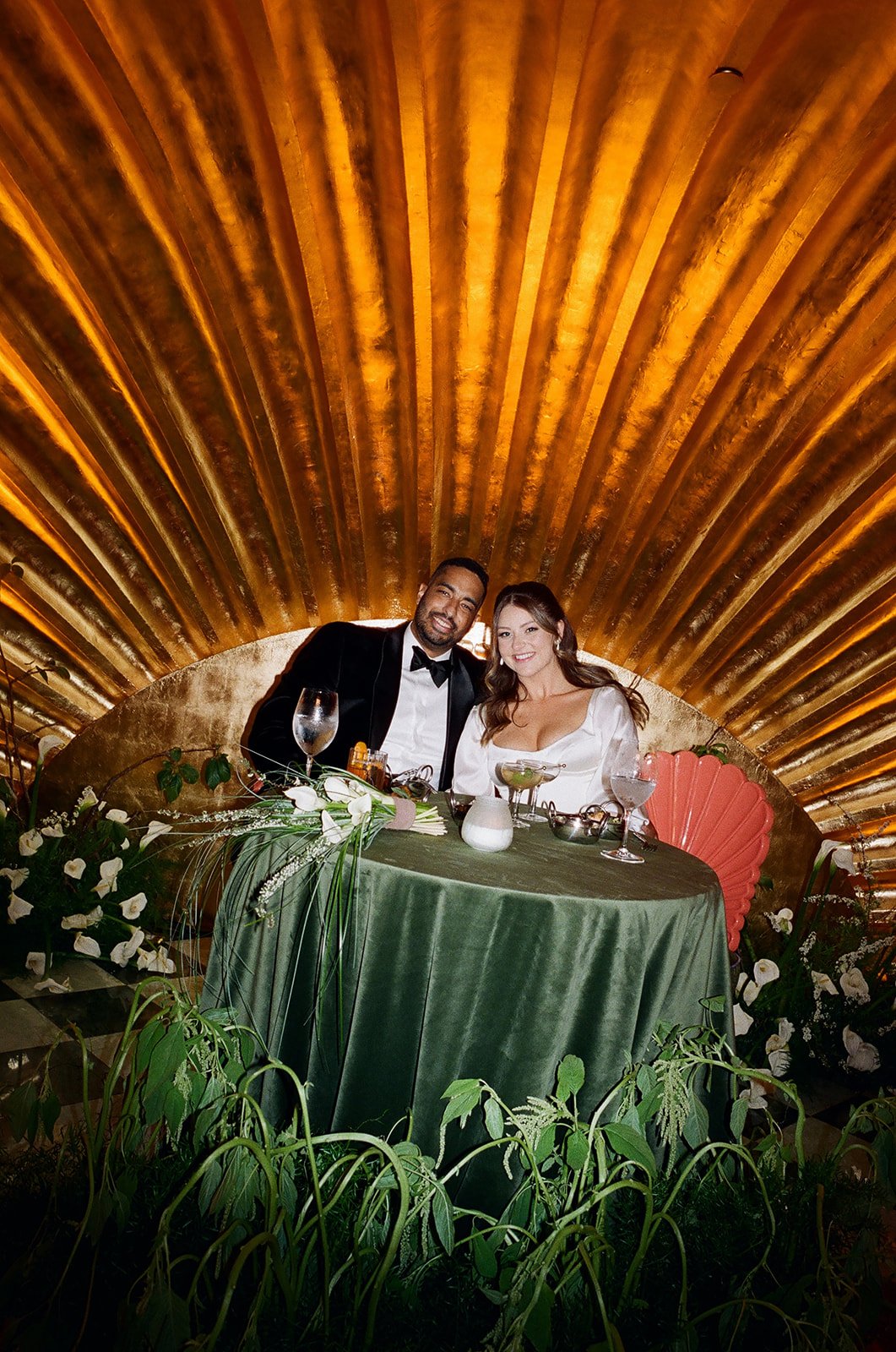 A newlywed couple dressed in wedding attire sits at a decorated table during their wedding reception, smiling happily.