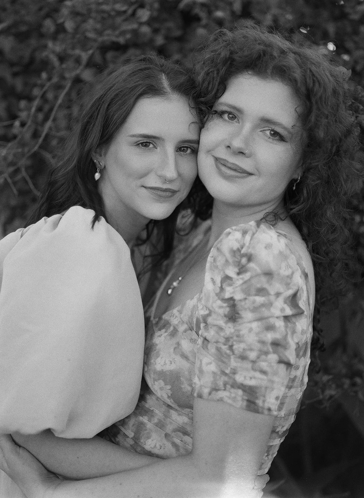 Two women hugging outdoors, smiling at the camera, with blurred natural background.