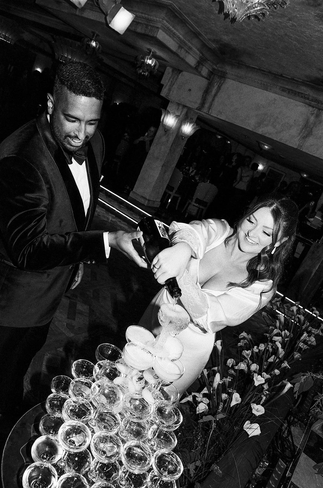 A woman in a white dress and a man in a black tuxedo pouring champagne into a tower of glasses at a celebration or wedding reception.