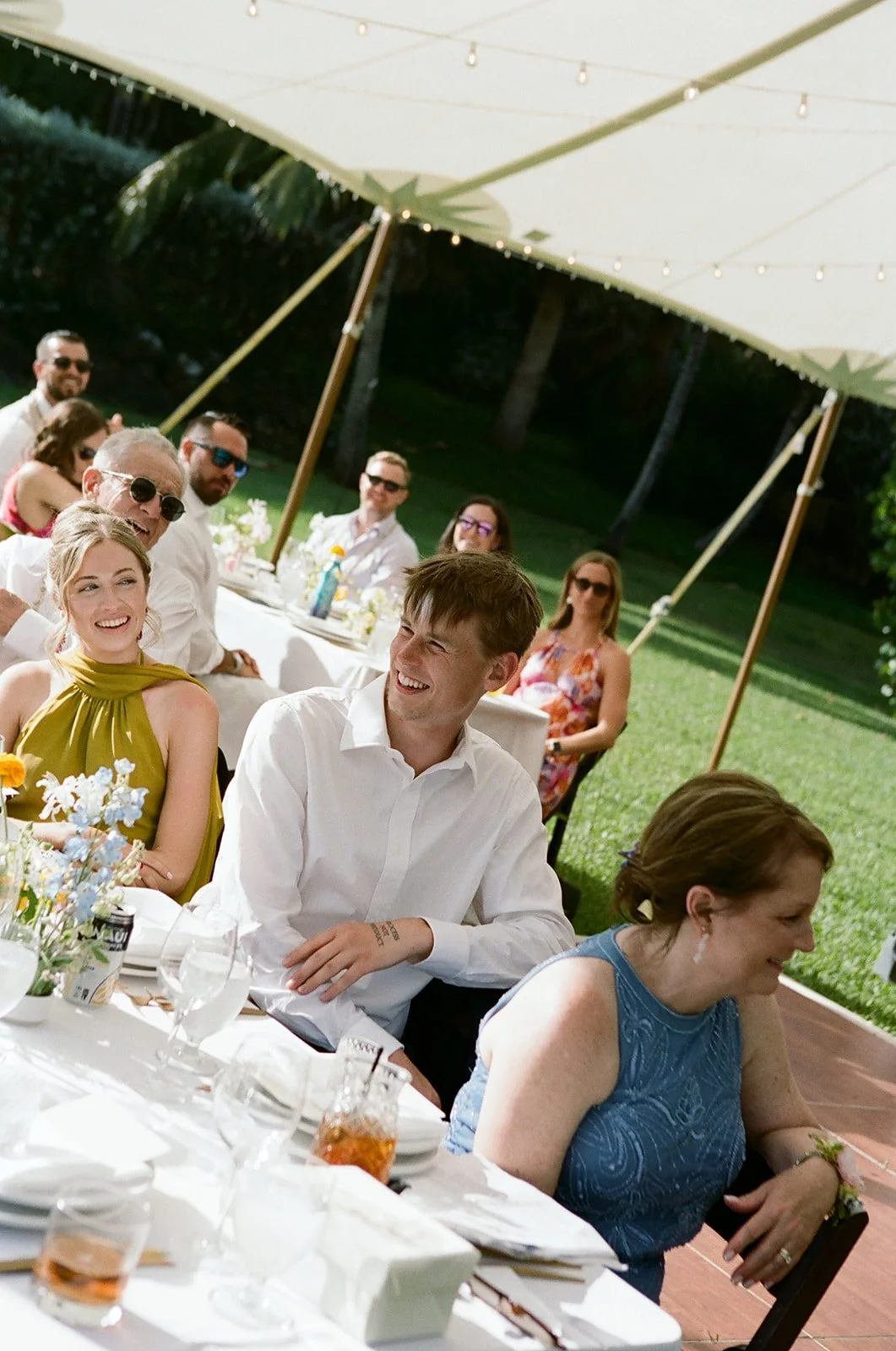 People sitting at a long outdoor event table under a large white canopy, enjoying a sunny daytime gathering.