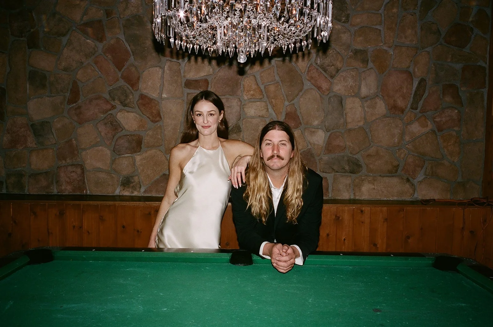 A woman in a white satin dress standing next to a man with long hair and a mustache, seated at a pool table, in a room with a stone wall and a large crystal chandelier hanging above.