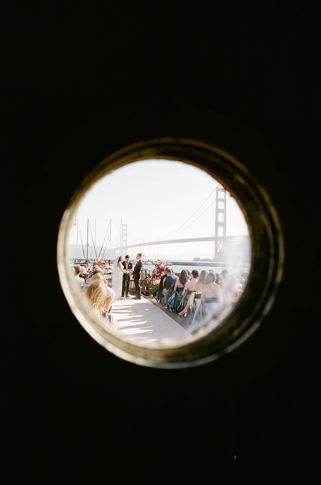 Wedding ceremony with bride and groom, guests seated outdoors near water, Golden Gate Bridge in the background, seen through a circular frame.