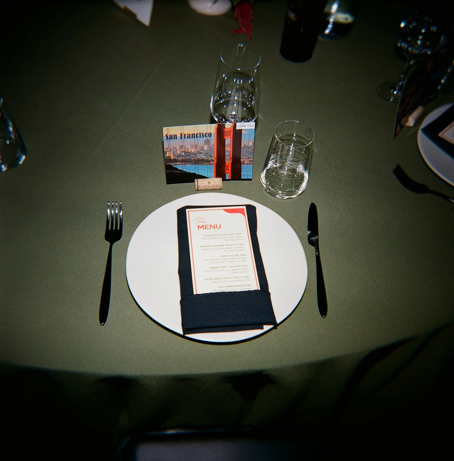 Table setting with a white plate, black napkin with a menu on top, two glasses, a fork on the left, a knife on the right, and a small card depicting the Golden Gate Bridge and skyline of San Francisco.