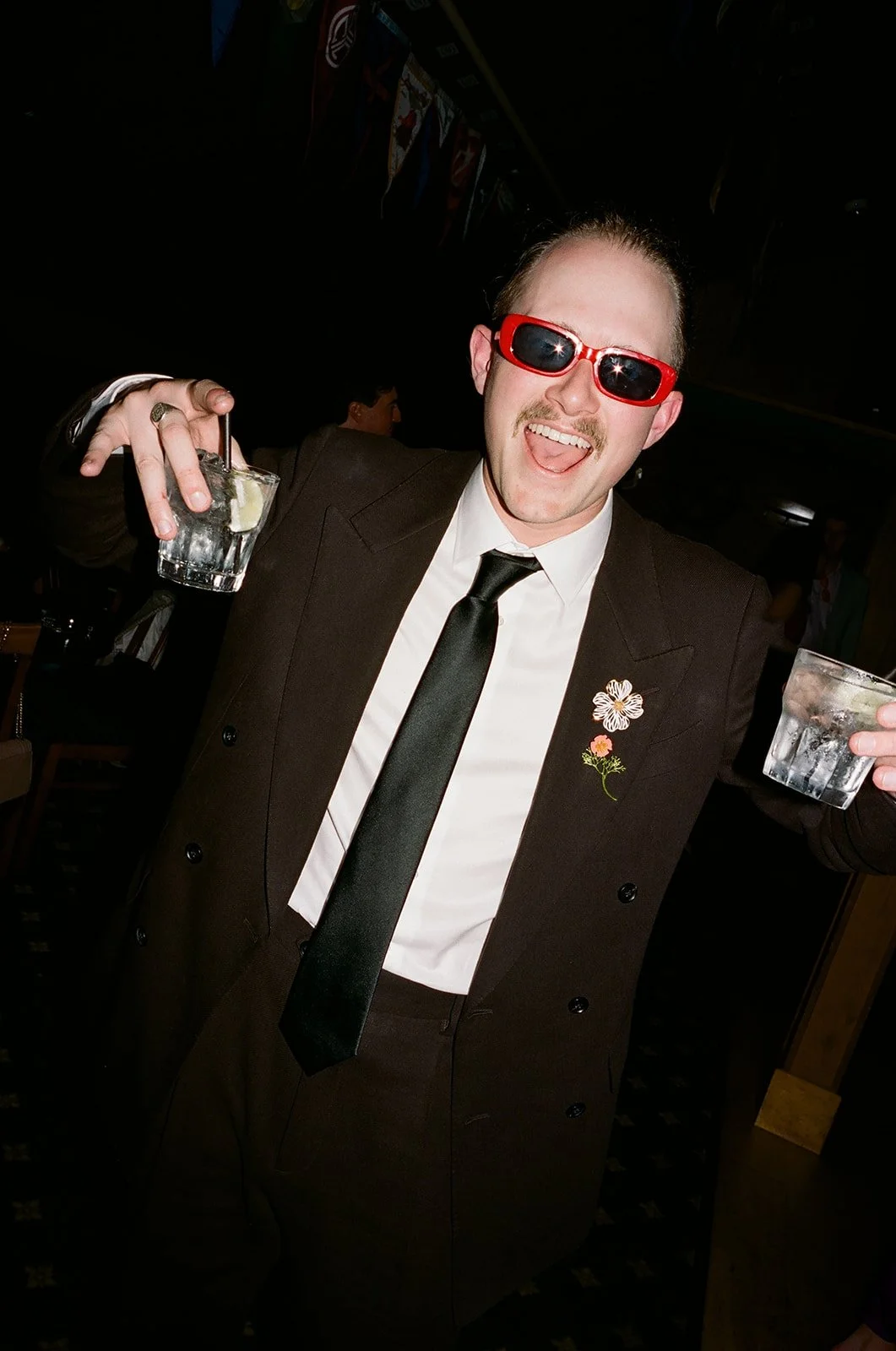 A man in a brown suit and tie, wearing red sunglasses, holding a glass of drink with ice and lemon, smiling, with a flower pin on his lapel, at a party or celebration.