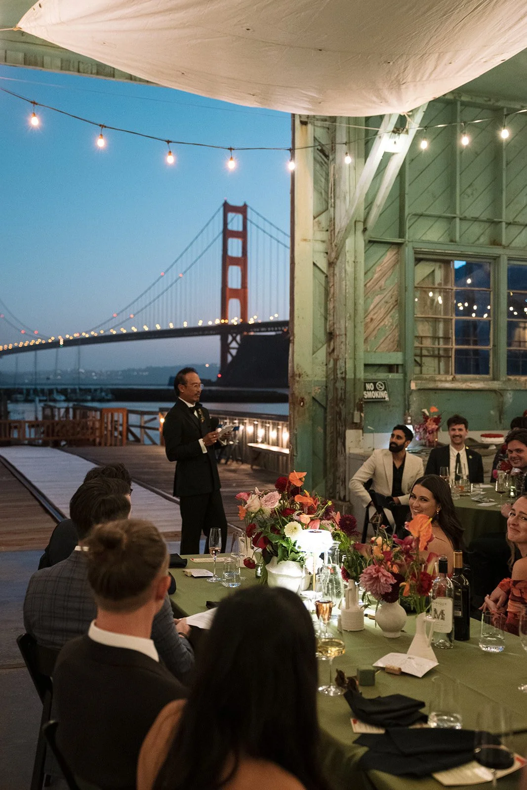 A dinner party with guests seated at a long table decorated with flowers, with the Golden Gate Bridge visible in the background. A man in a tuxedo is speaking to the seated guests, under string lights in an outdoor setting at dusk.