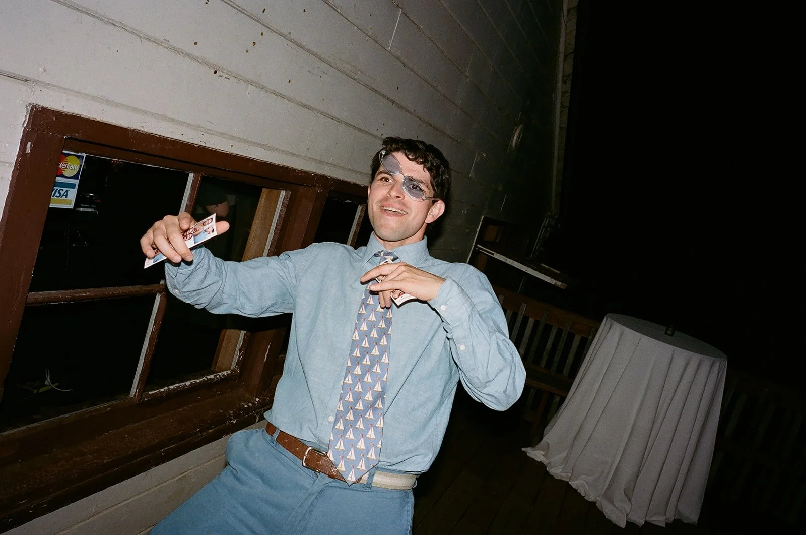 A young man in a dress shirt, patterned tie, and glasses is smiling and dancing at a party, holding a piece of paper or card in one hand, with a window and a table covered with a white tablecloth in the background.