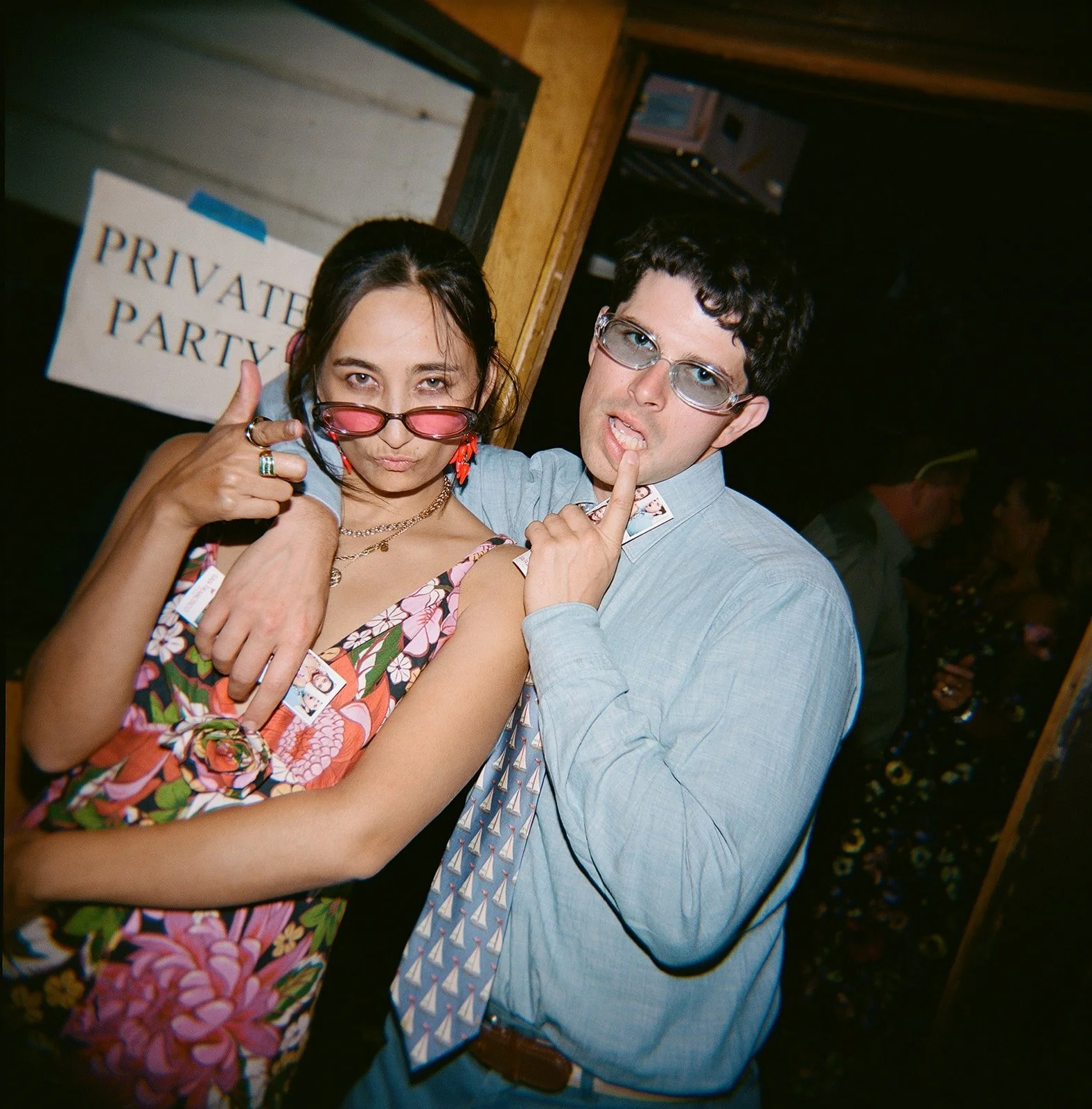 Two young adults, a woman and a man, posing playfully together at a social event, both wearing glasses and making expressive gestures near their faces.