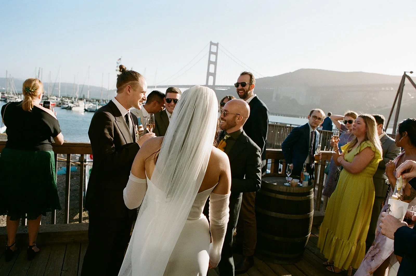 A wedding reception outdoors near San Francisco, with the Golden Gate Bridge in the background, featuring guests in formal attire enjoying drinks and conversation.