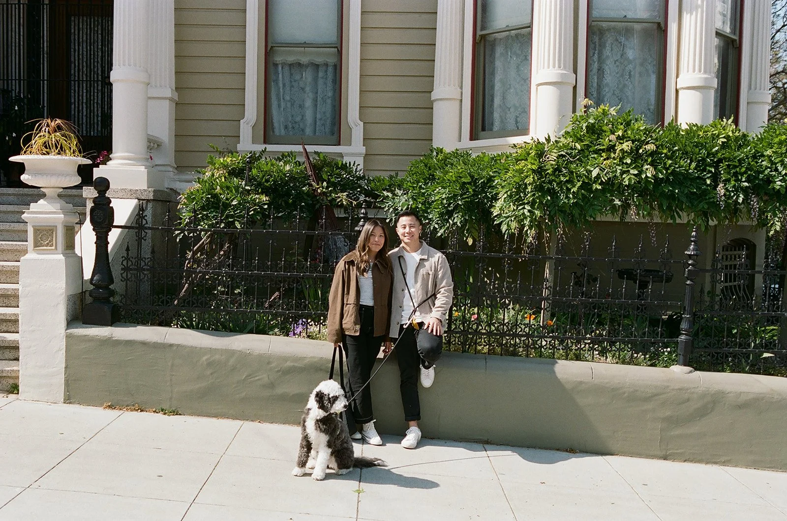 A young couple standing on a city sidewalk with a dog, in front of a residential house with lush greenery and colonial-style architecture.