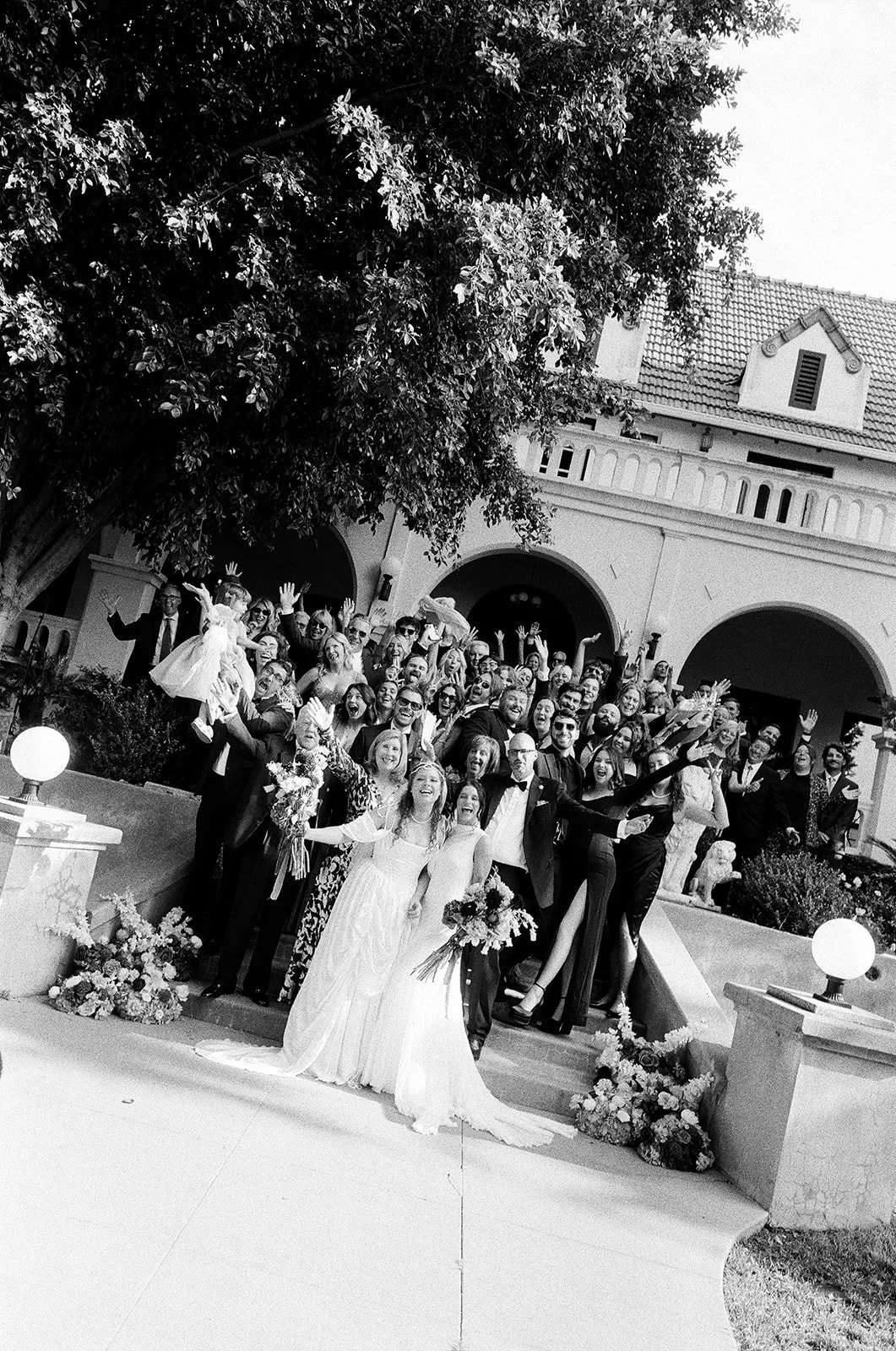 Black and white photo of a large group of people celebrating a wedding in front of a house with a tiled roof and large arches, including the bride and groom in wedding dresses and tuxedos, with floral arrangements around the stairs.