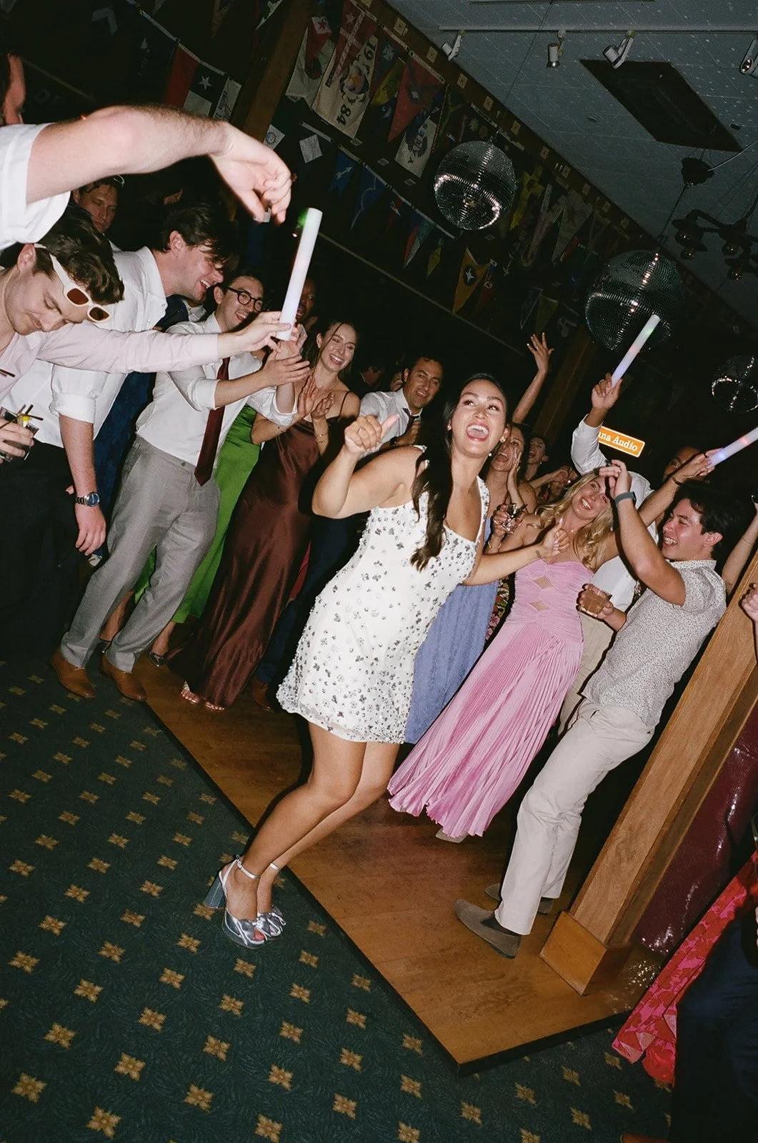 A group of people celebrating and dancing at a party or wedding reception, with some holding glow sticks, in a decorated venue with flags and disco balls.