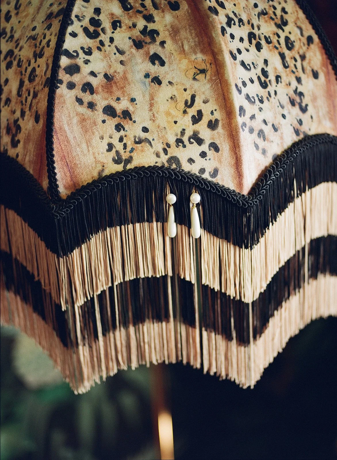 Close-up of a decorative lamp with a leopard print fabric shade, black fringe and beads, and a wooden fringed trim.