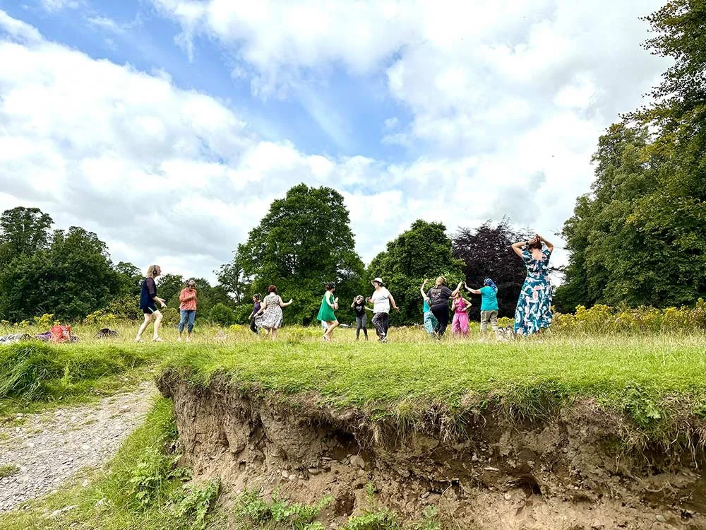 A group of ladies dancing on a riverbank surrounded by trees