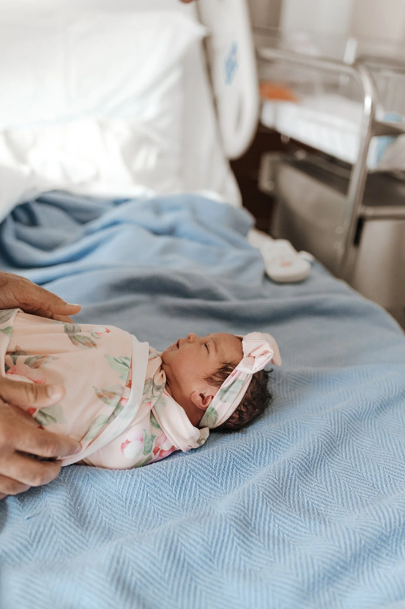 A newborn baby girl lying on a hospital bed, wearing a floral headband and outfit, with an adult's hand gently touching her side in a hospital room, at Centennial Hill Hospital.
