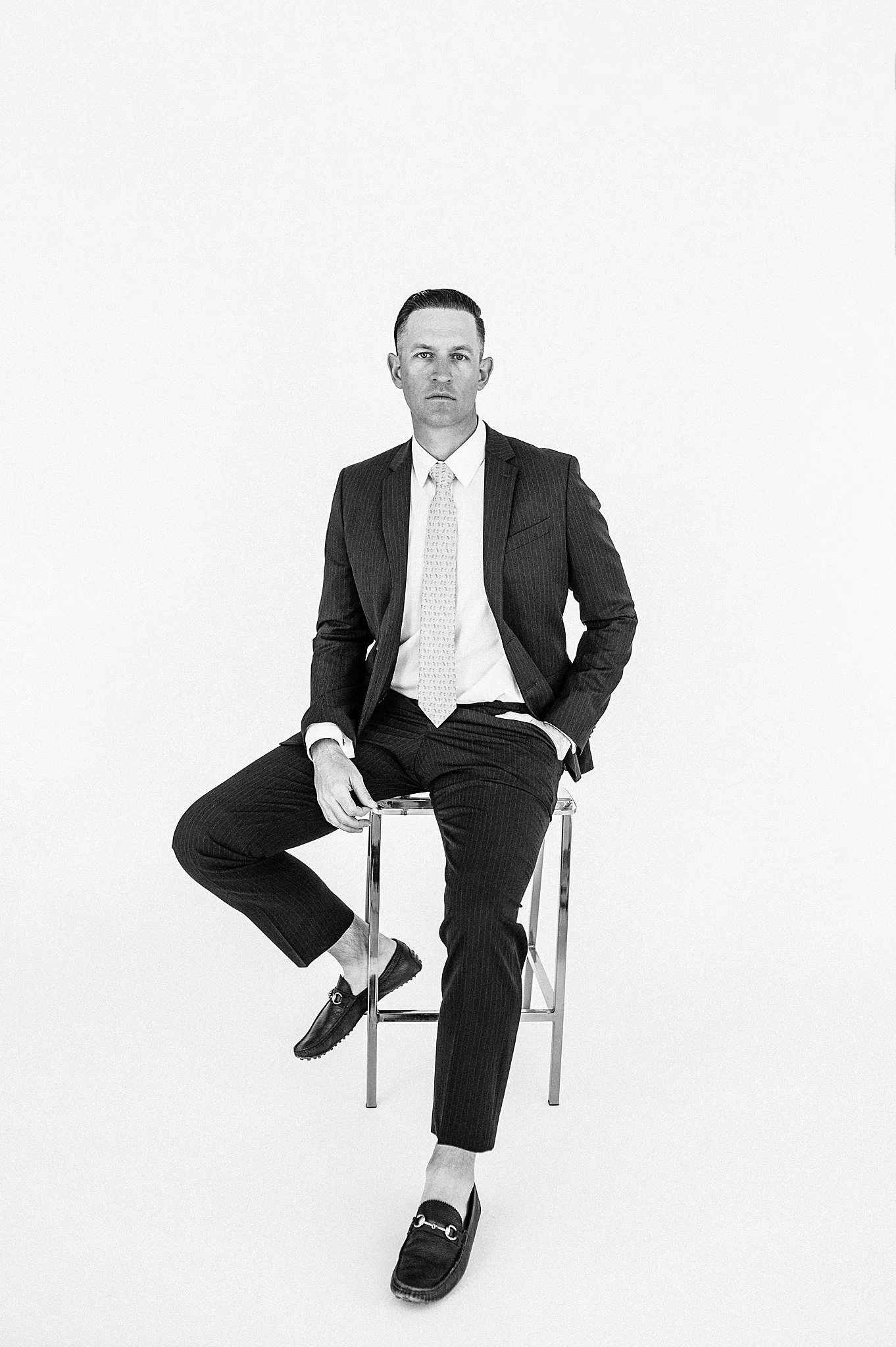 Black and white photo of a man in a business suit sitting on a stool against a plain background.