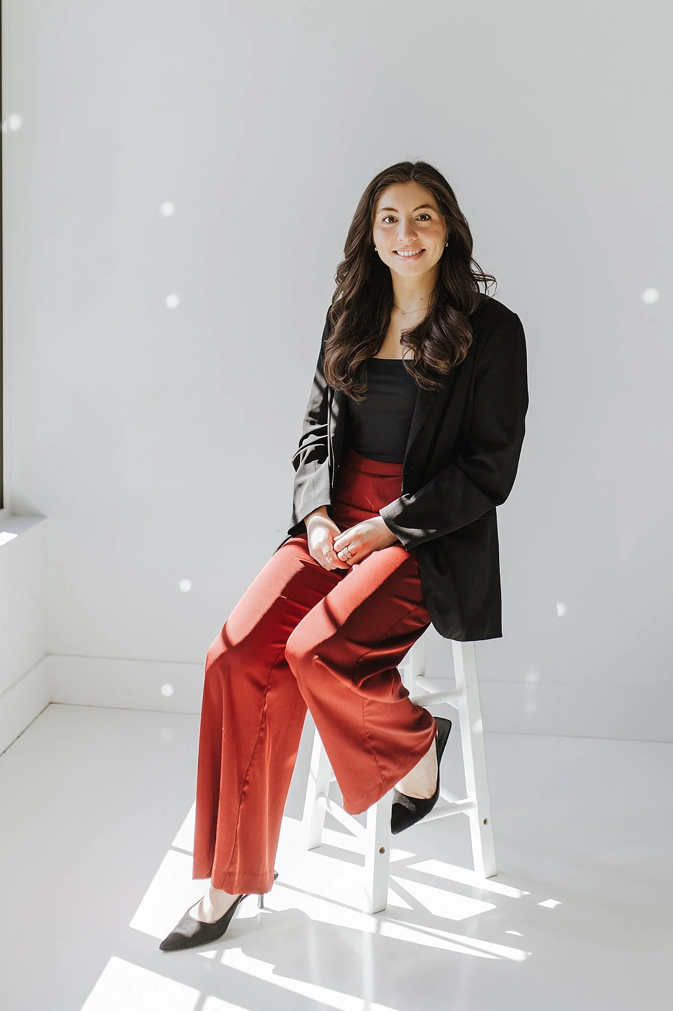 Young woman sitting on a white ladder in a bright studio with a white background and window, wearing a black blazer, black top, red wide-leg pants, and black high heels, smiling at the camera.
