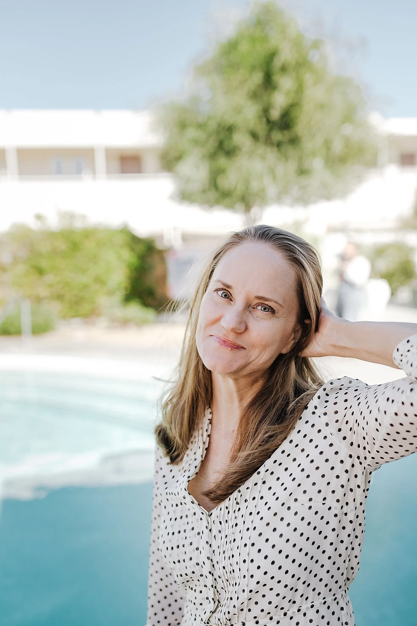 A woman with long brown hair and blue eyes, wearing a white blouse with black polka dots, standing outdoors near a pool with trees and a building in the background.