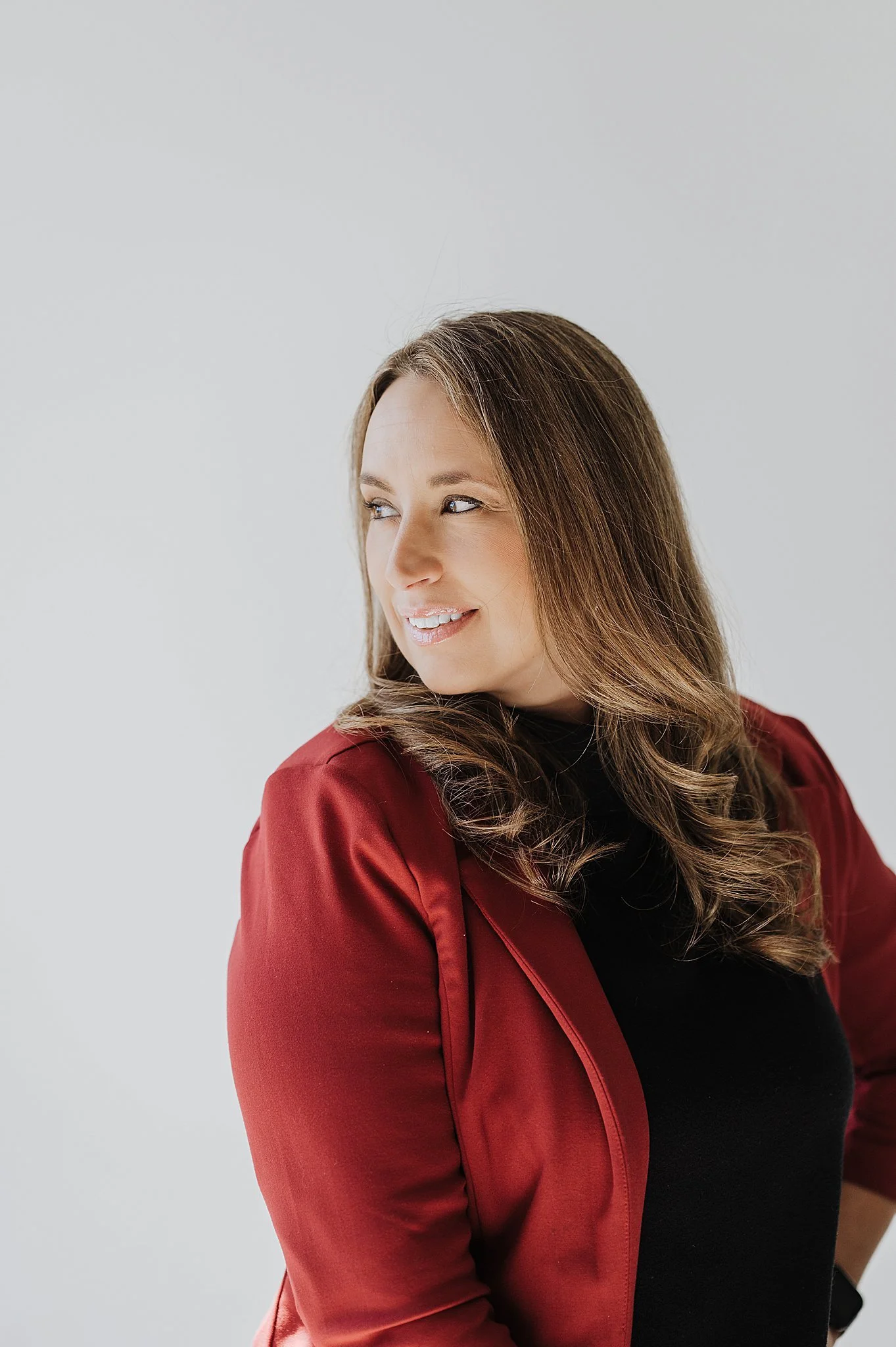 A woman with wavy brown hair wearing a black top and a red jacket, looking to her right with a slight smile, against a plain white background.
