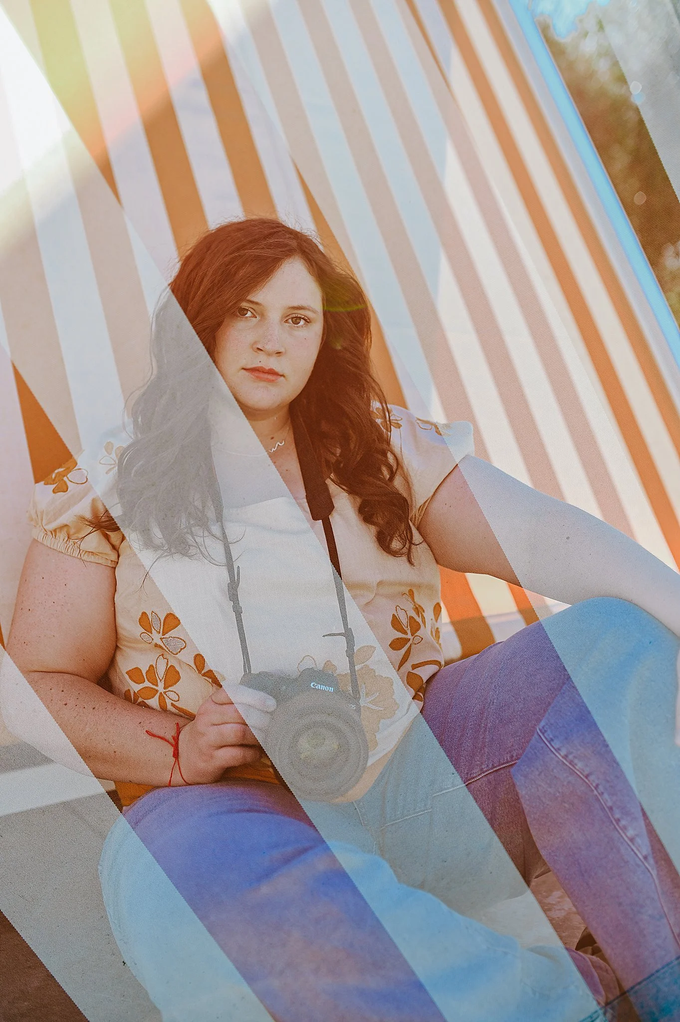 Young woman with red hair holding a Canon camera, sitting on a striped orange and white chair outdoors during sunset.