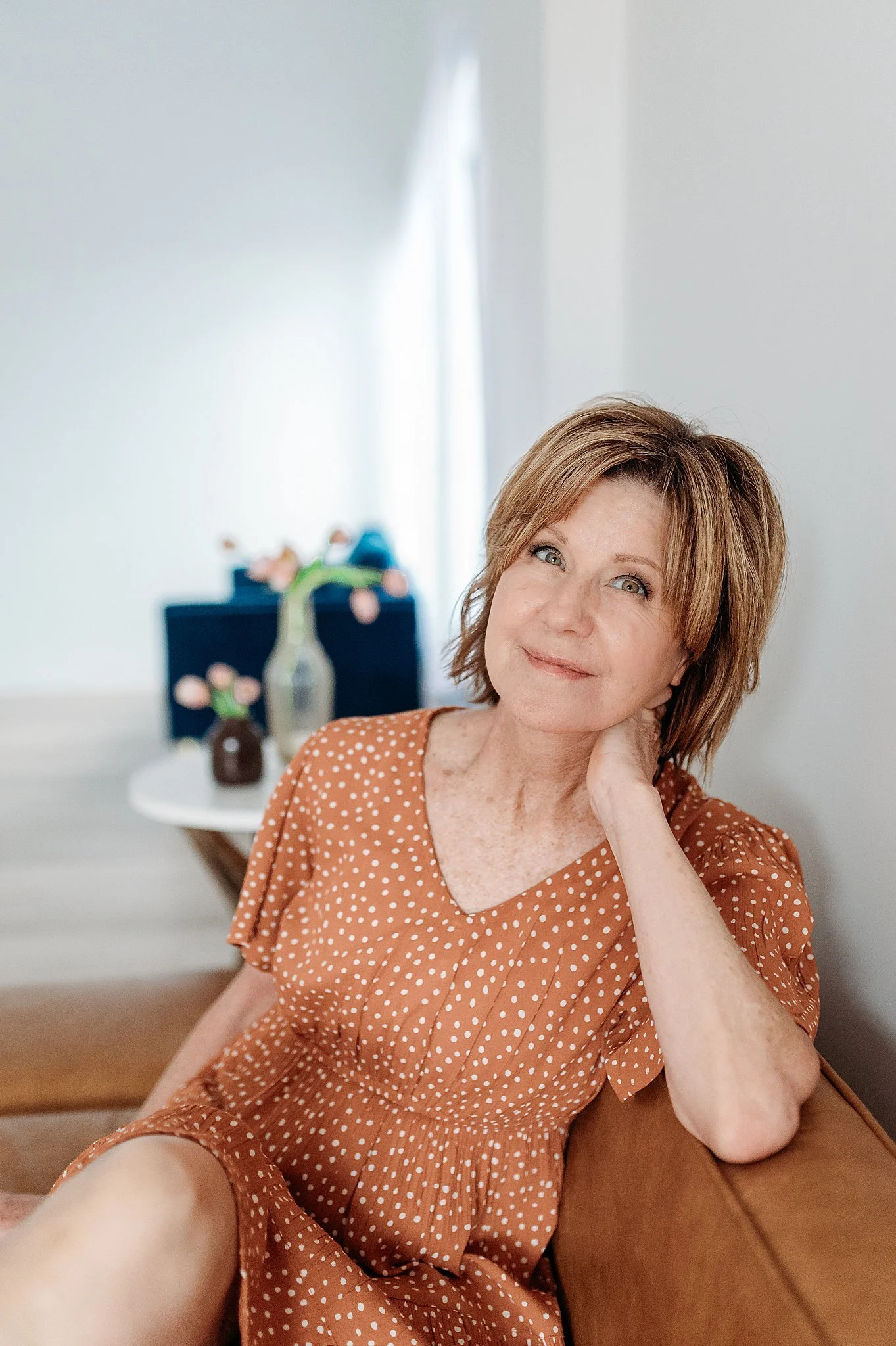 A woman with reddish-brown hair and blue eyes sits on a brown couch, resting her head on her hand and smiling softly. She wears a rust-colored dress with white polka dots. In the background, there is a small round table with a vase of pink flowers an